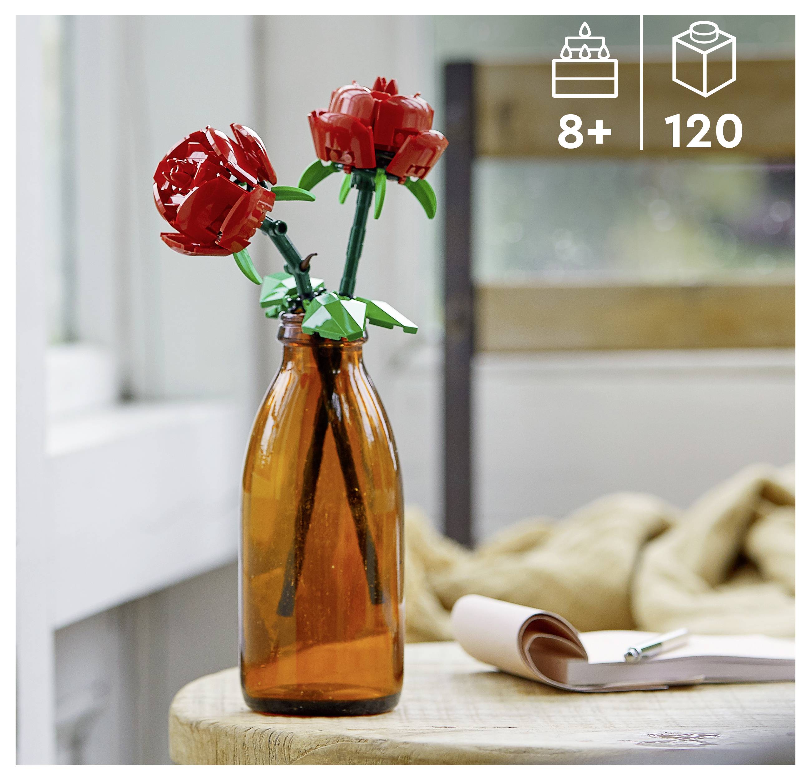 Een foto van een bruine glazen fles op een tafel, met twee rode LEGO-rozen met groene bladeren. Heldere kamer met onscherpe achtergrond.