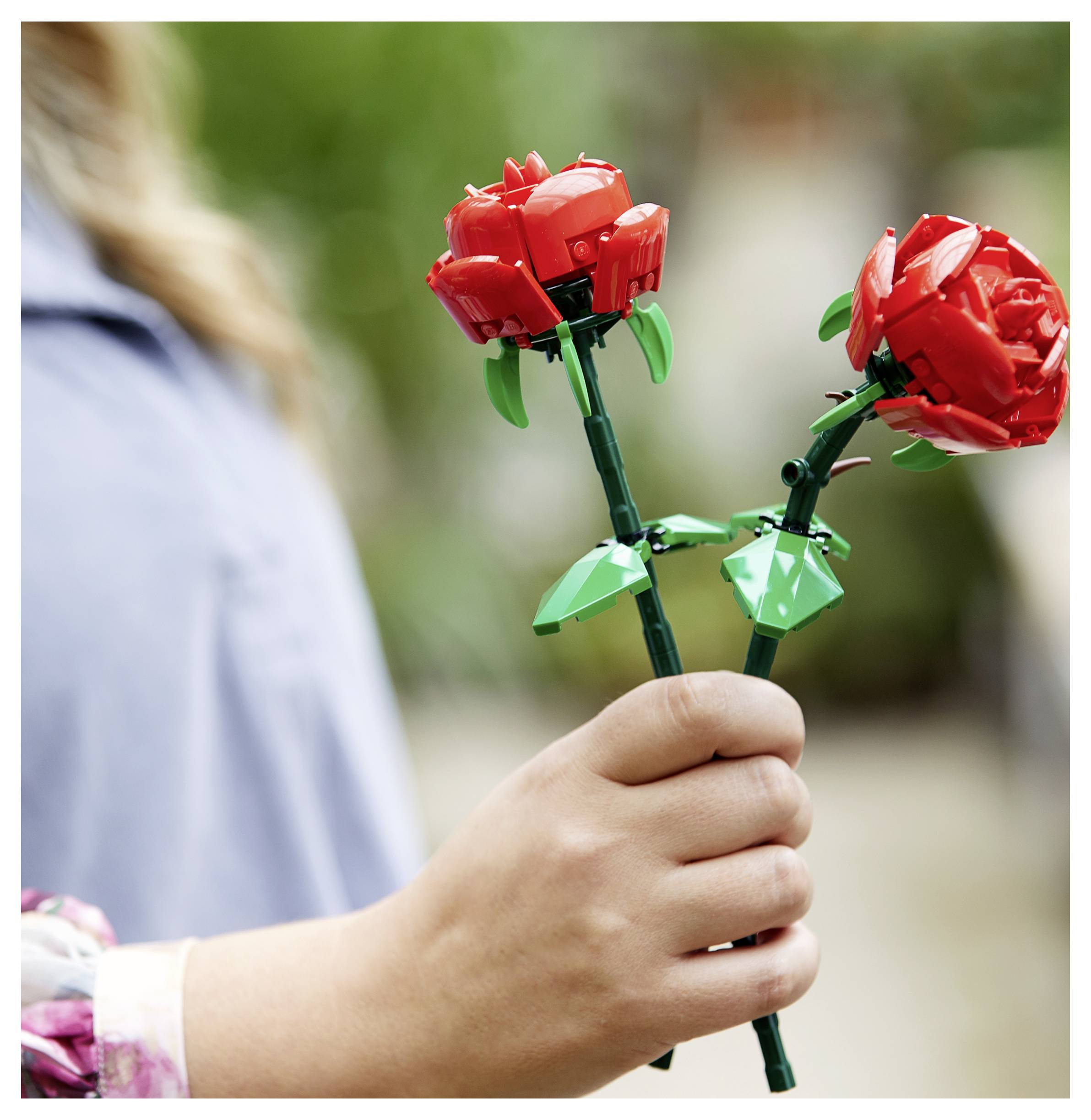 Een persoon die twee rode Lego-rozen vasthoudt, één in elke hand, in een buitenomgeving.