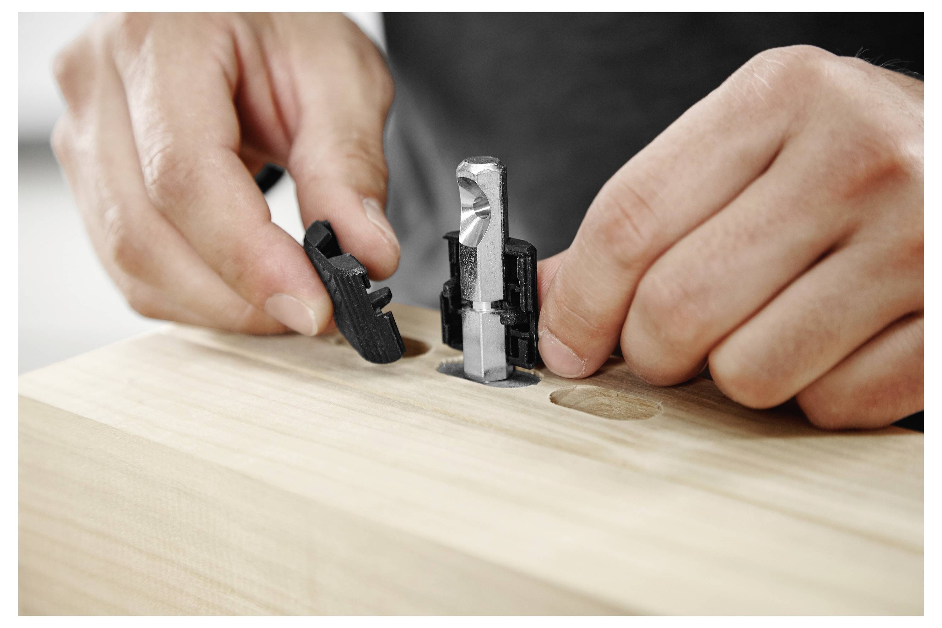 Handen die een zwart en zilver hardwareconnector in een houten oppervlak met voorgeboorde gaten samenstellen, gericht op constructiedetail.