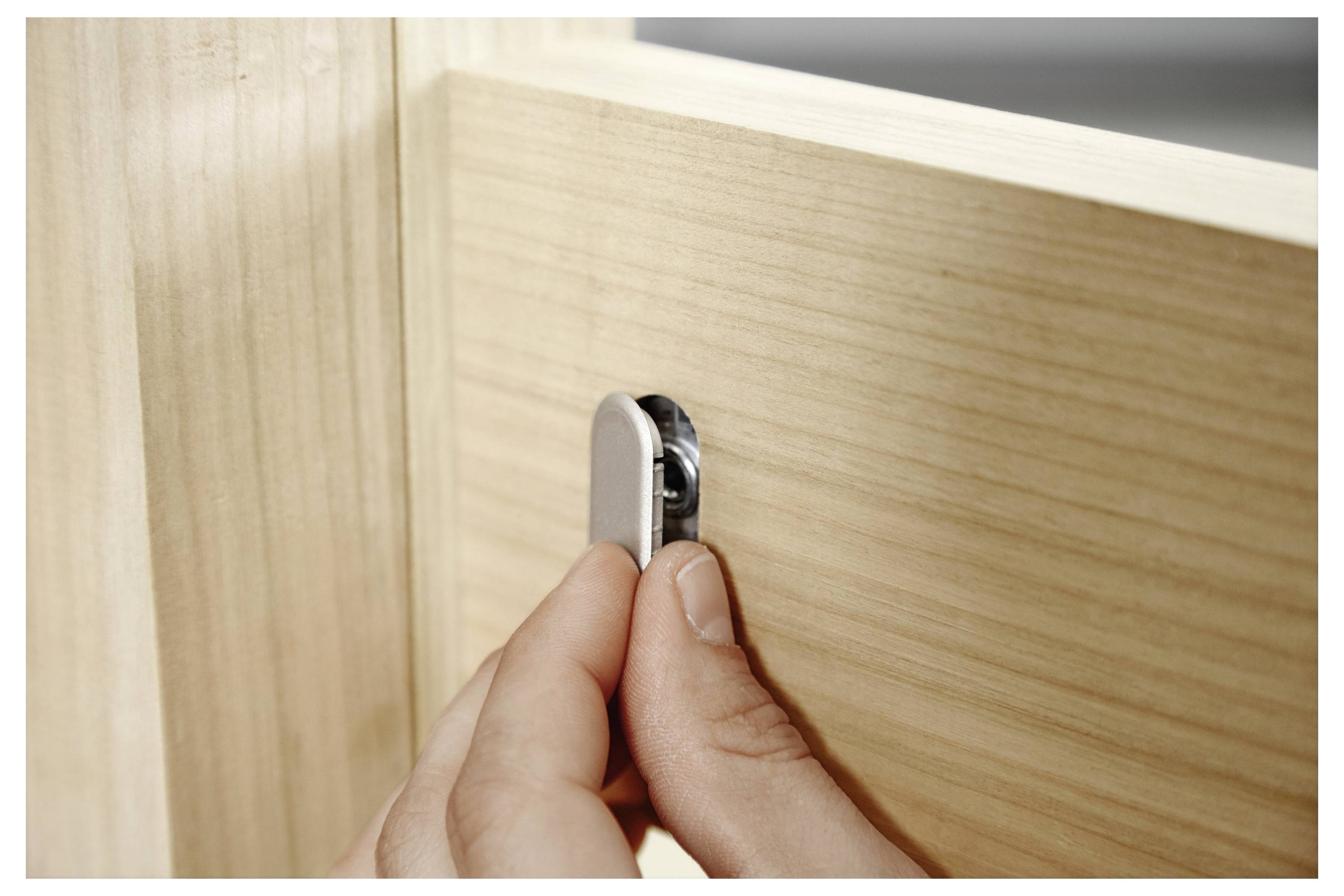 Een hand stelt een metalen nockslot op een houten meubelstuk in, wat op montage of installatie wijst.