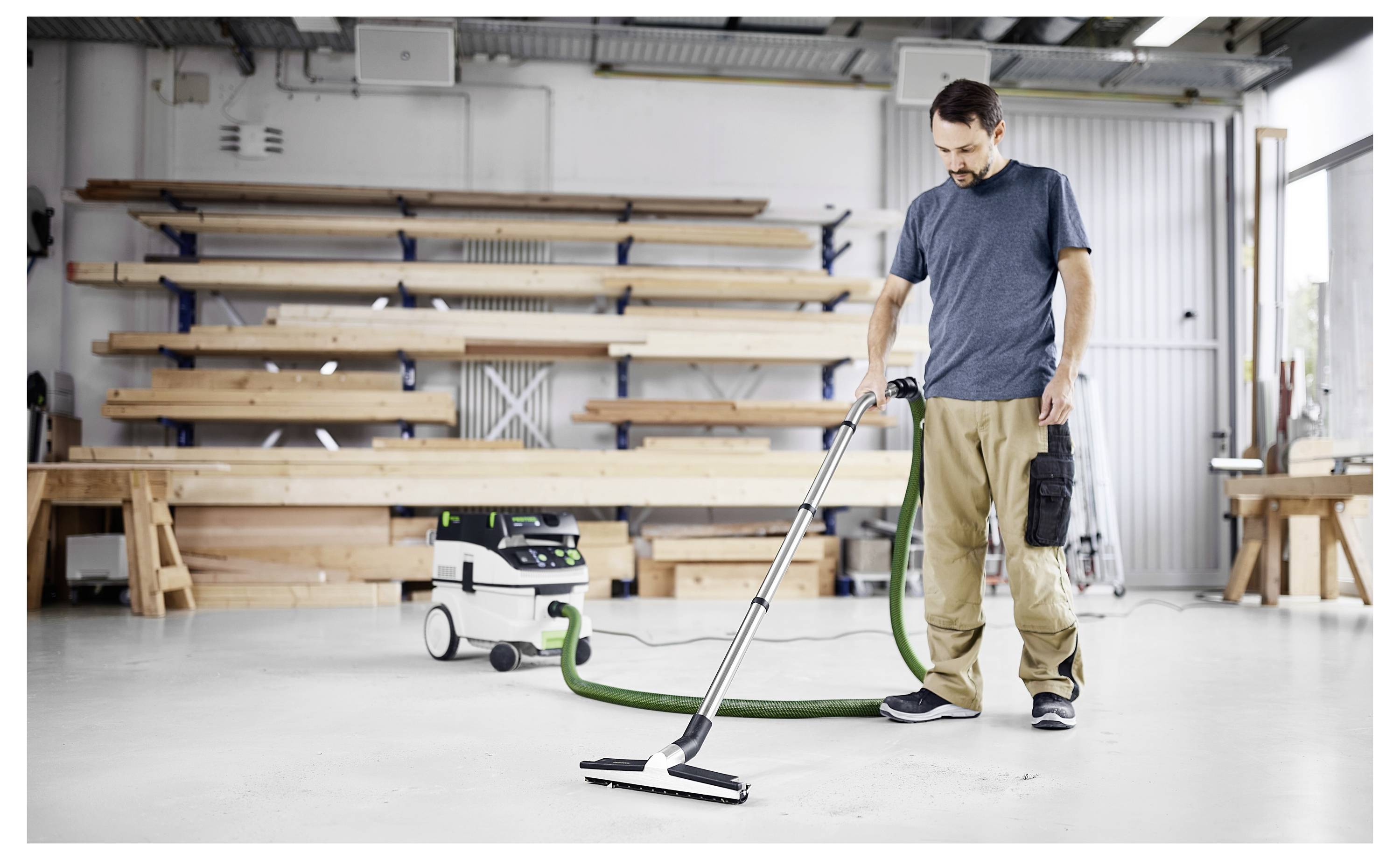 Een persoon die een stofzuiger op de vloer gebruikt in een werkplaats met houten planken op schappen op de achtergrond, wat schoonmaak- of onderhoudswerkzaamheden aantoont.