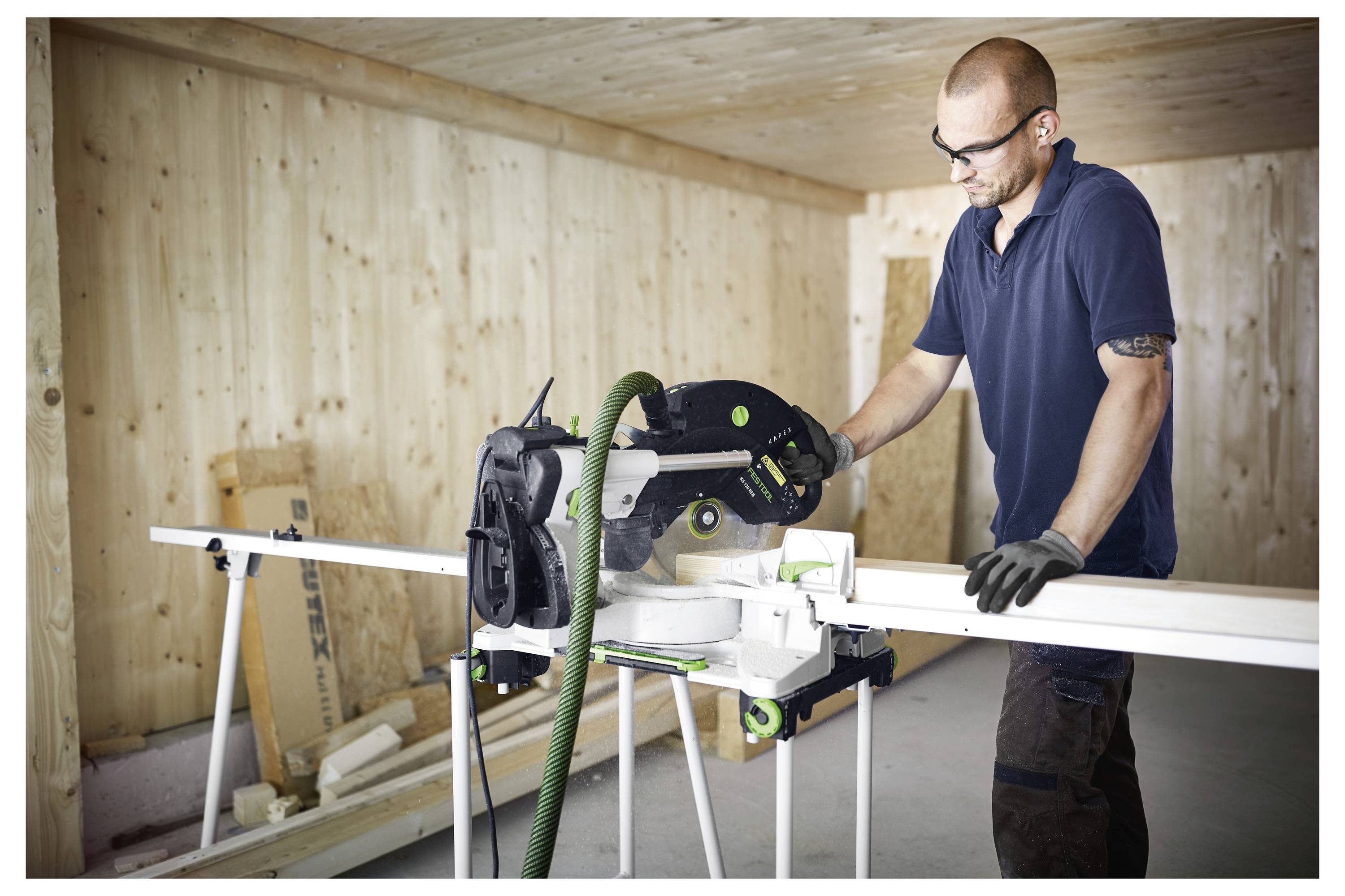 Een persoon in veiligheidsuitrusting bedient een tafelzaag in een houten werkplaats, waarbij een plank wordt gesneden, met nadruk op houtbewerking en precisie.