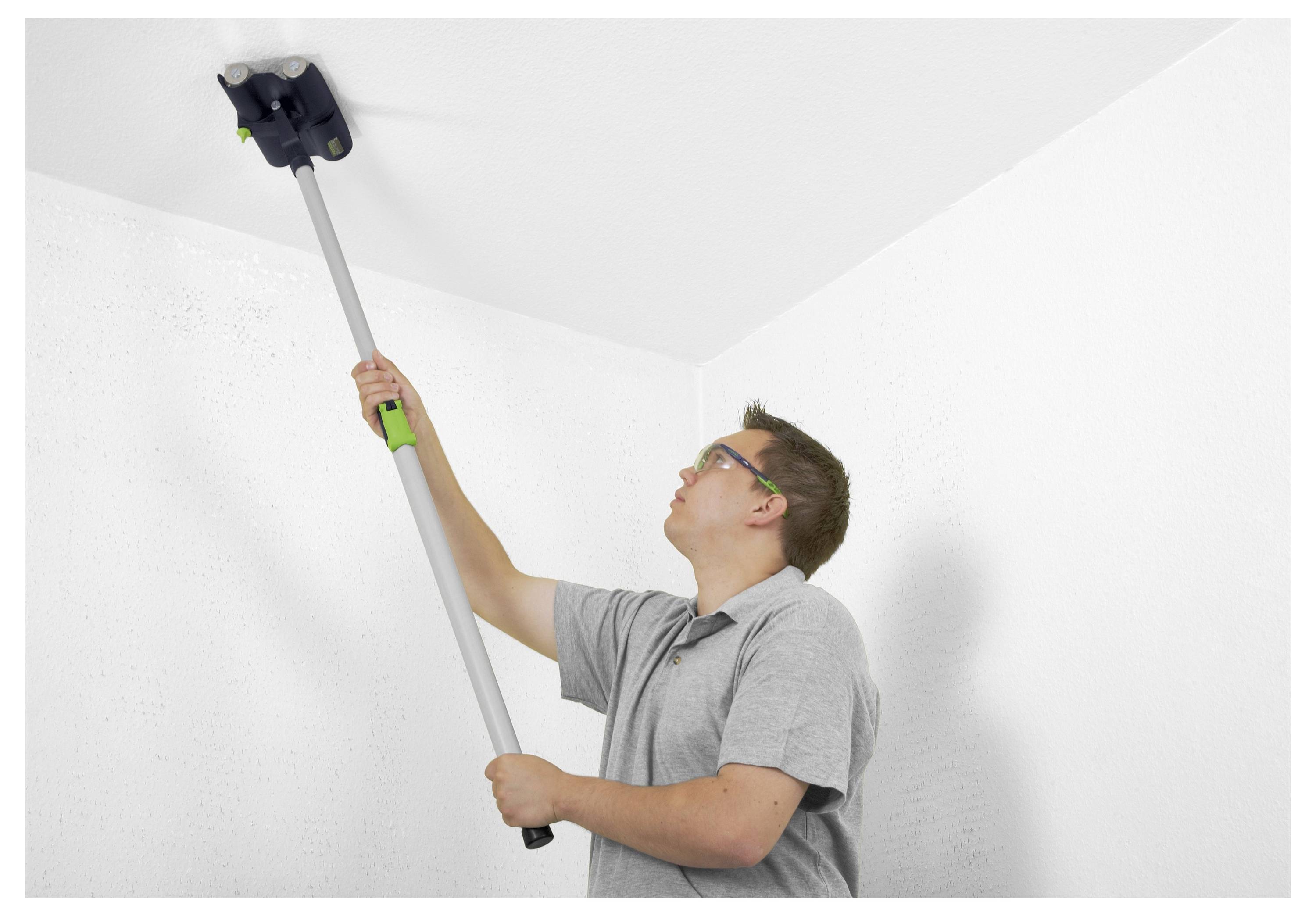 Een persoon met een bril gebruikt een gereedschap met lange steel om een wit plafond schoon te maken, gekleed in een lichtgrijs shirt, en concentreert zich op een hoekgebied.