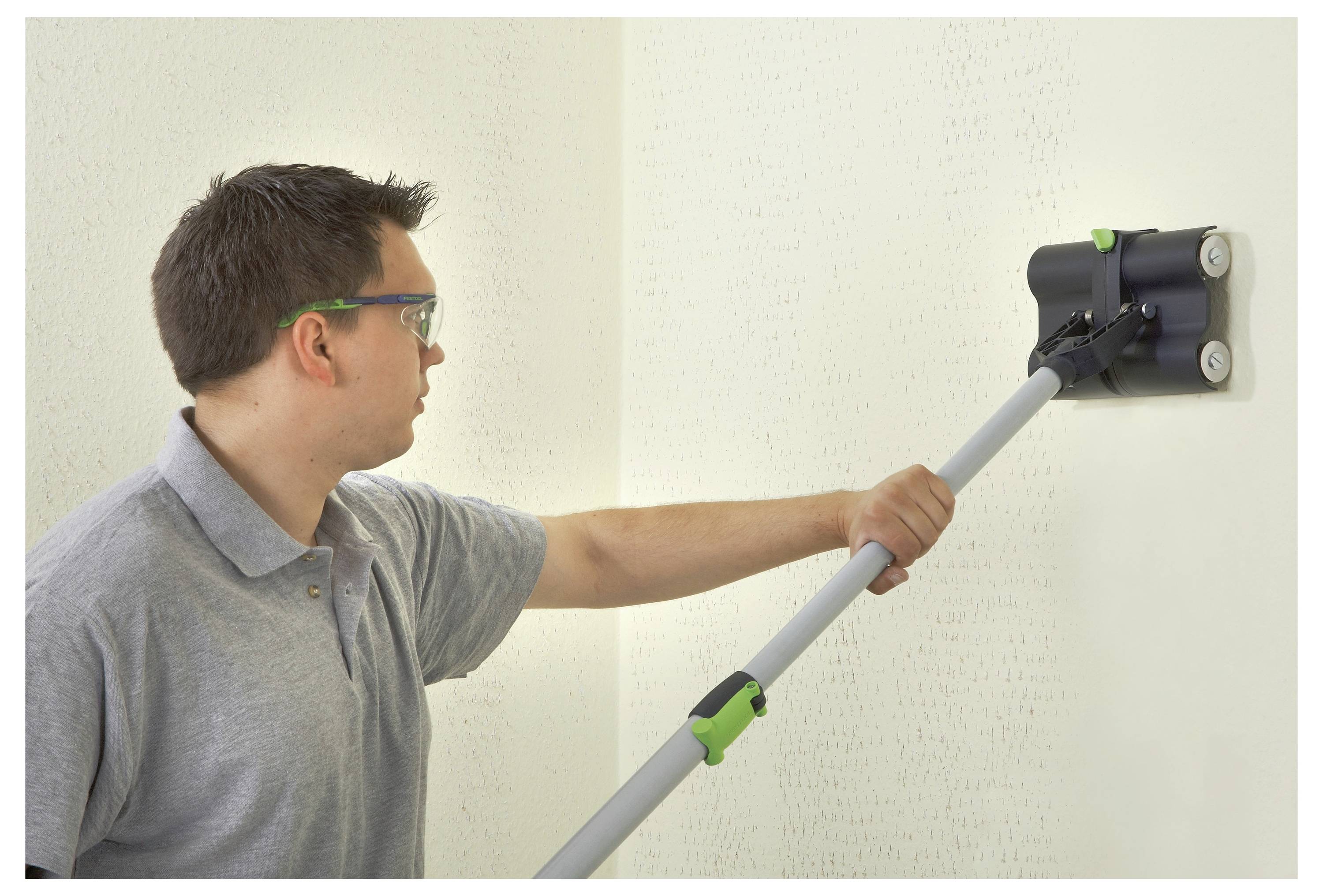 Een persoon die een veiligheidsbril draagt, gladstrijkt een muur met een lang handvat slijptool.