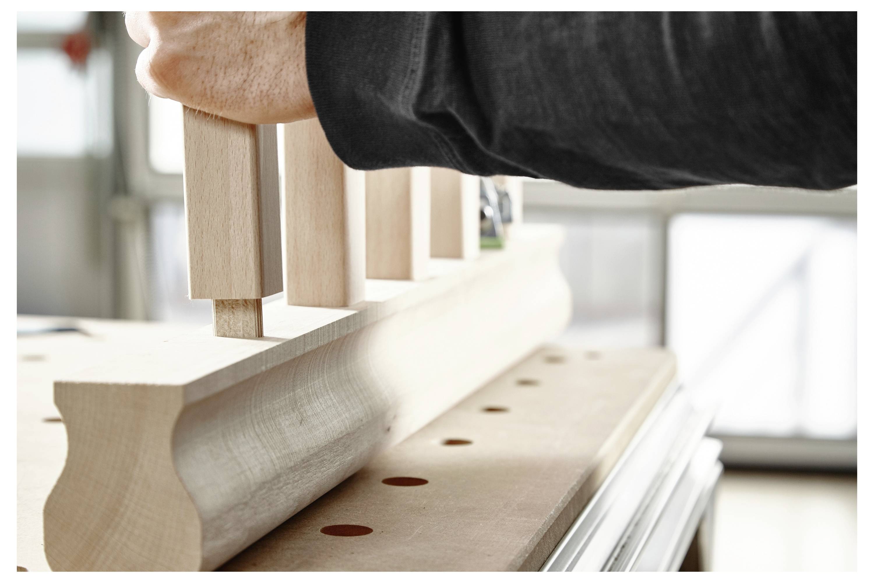 Een persoon steekt houten pennen in een stuk gegroefd hout op een werkbank, wat suggereert dat meubels of kasten worden samengesteld.