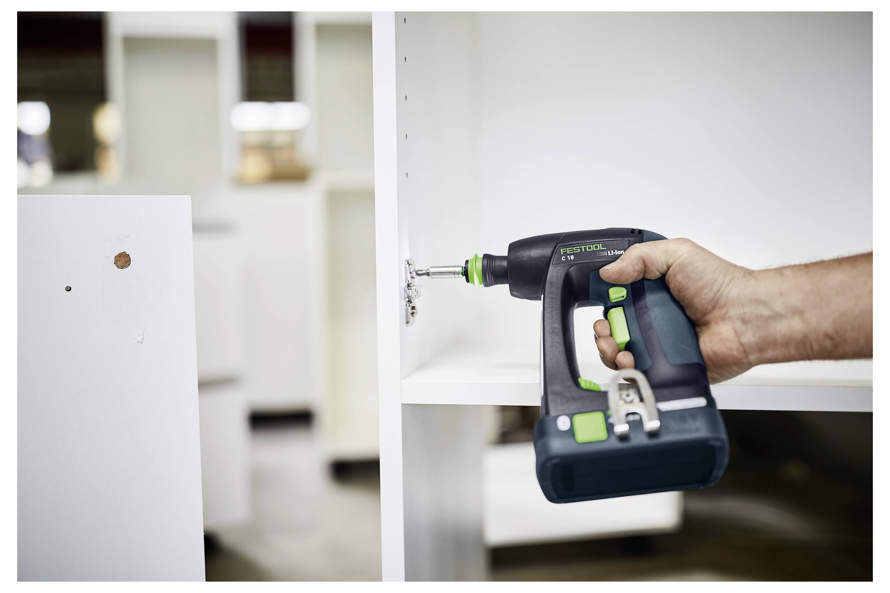 Een persoon gebruikt een elektrische schroevendraaier om een witte kast in elkaar te zetten. De boor is groen en zwart, en de omgeving lijkt een werkplaats te zijn.