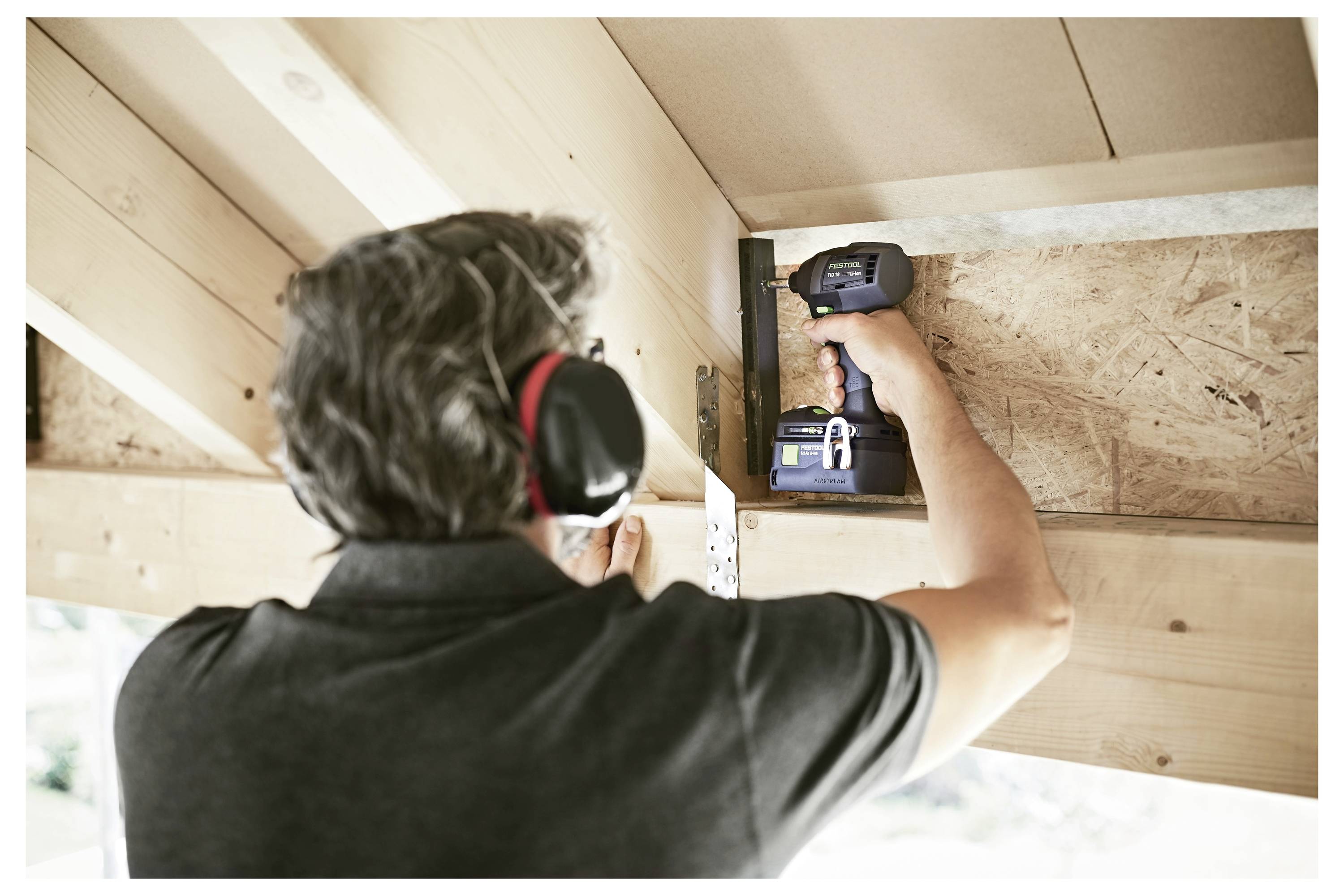 Een persoon die oorbescherming draagt, gebruikt een boormachine om een houten balk onder een plafond vast te zetten, wat wijst op bouw- of renovatiewerkzaamheden.