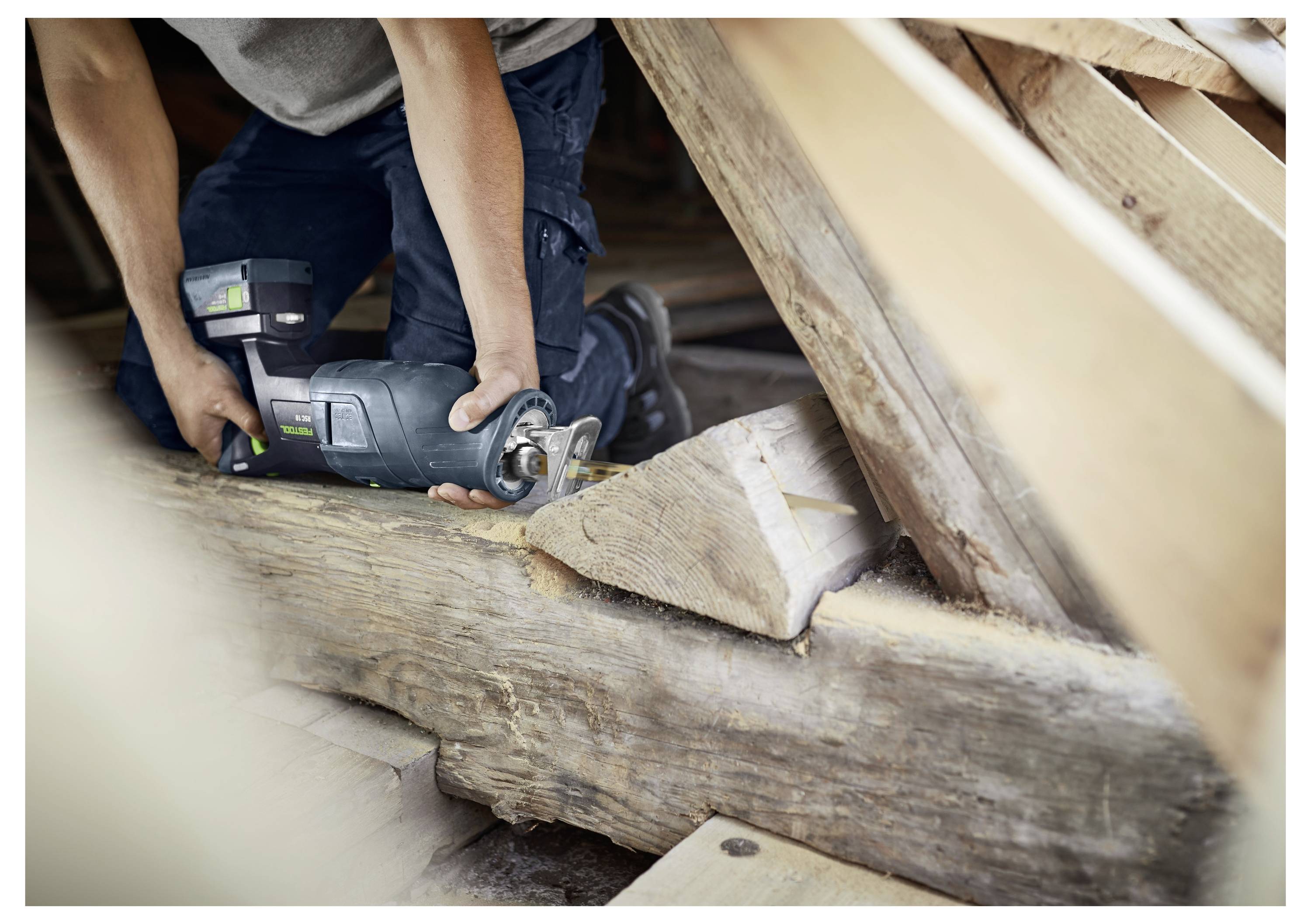 Een persoon gebruikt een cirkelzaag om een houten balk in een zolderruimte met zichtbare dakspanten door te zagen, wat wijst op bouw- of renovatiewerkzaamheden.