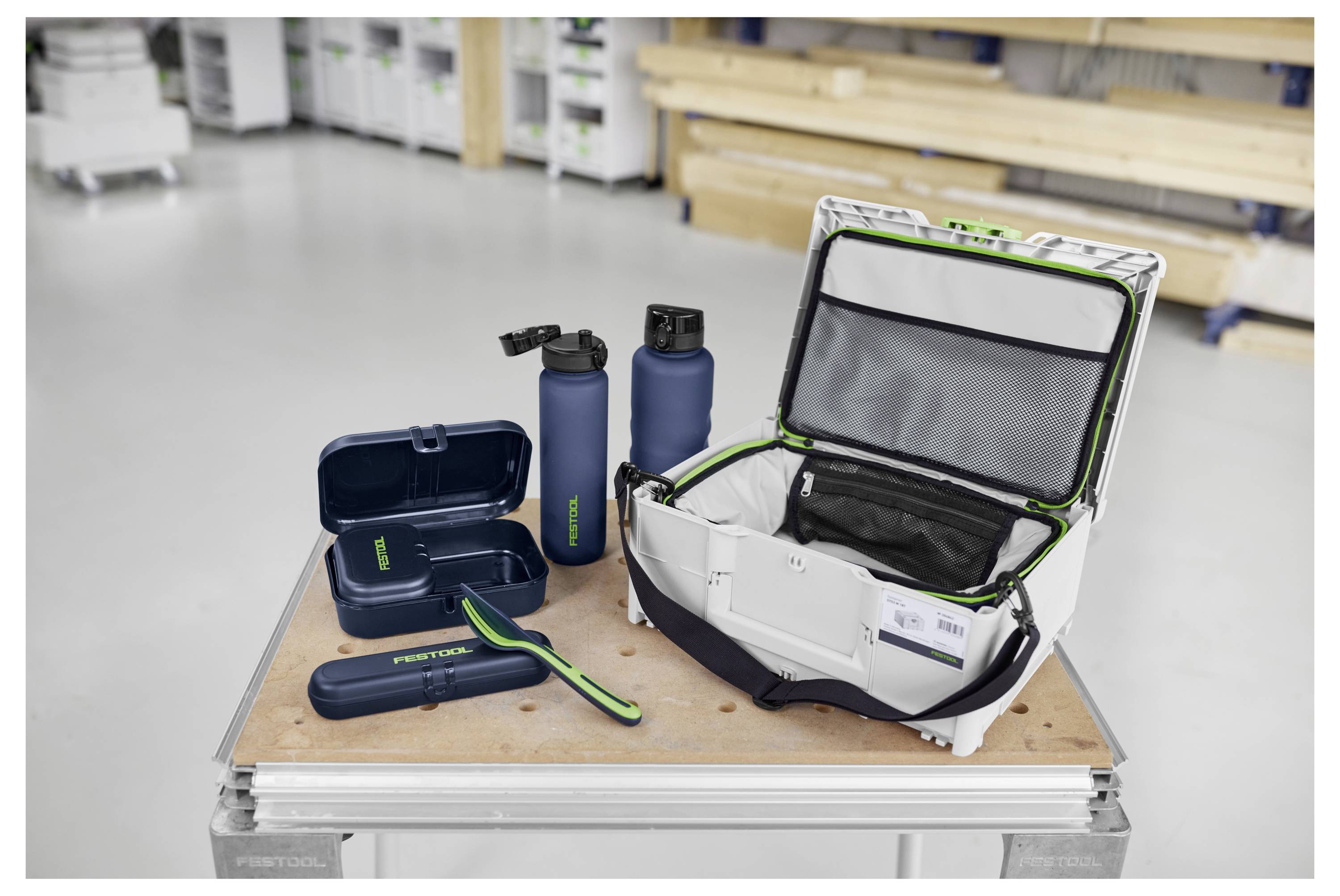 "Festool" merkt lunchbox, drinkflessen en tas weergegeven op een houten werkbank in een werkplaatsomgeving met gereedschap op de achtergrond.