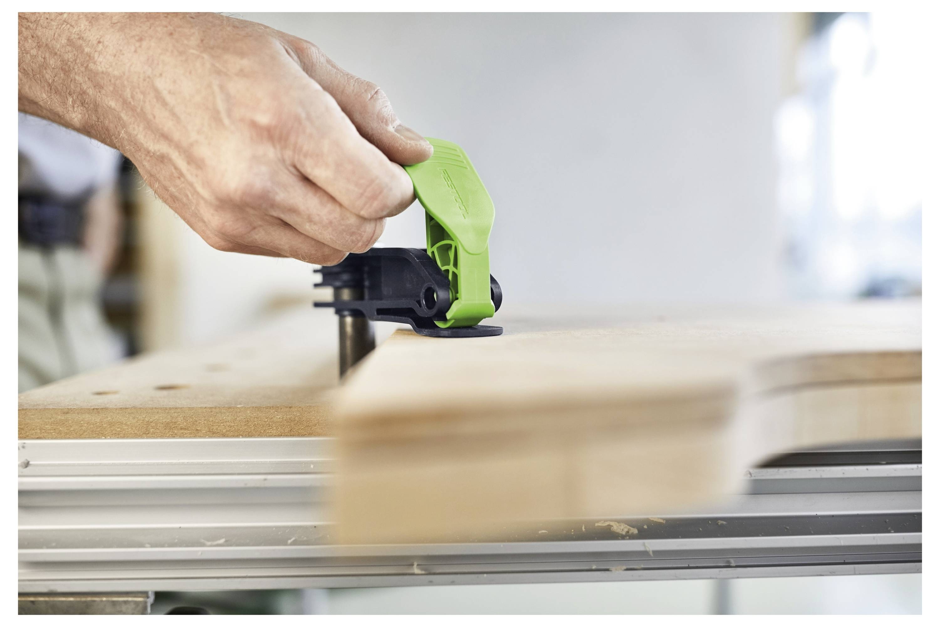 Een hand gebruikt een groen gereedschap om een houten plank stevig aan een werkbank vast te klemmen, wat een houtbewerkingsproces in uitvoering illustreert.