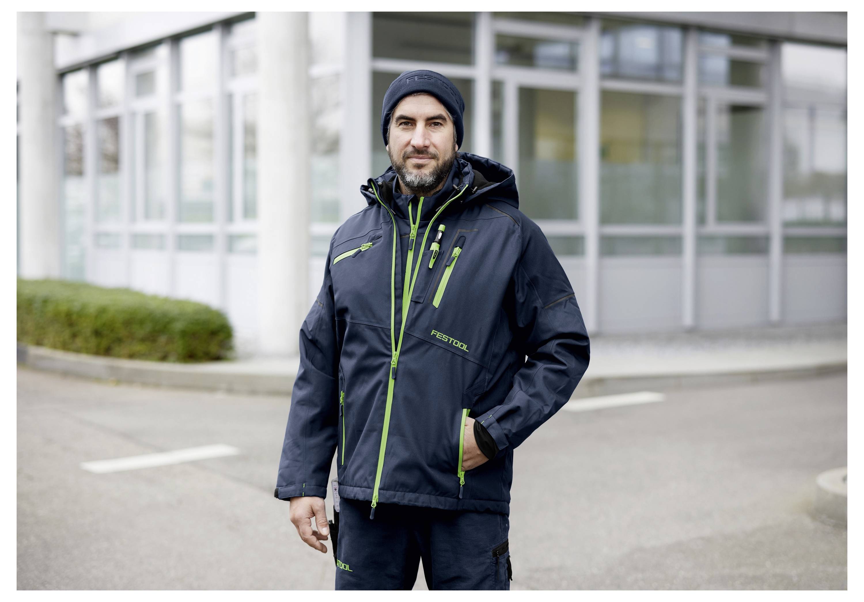 Een persoon in winterkleding staat buiten voor een modern gebouw, gekleed in een donker jasje, muts en broek, met handen in zakken.