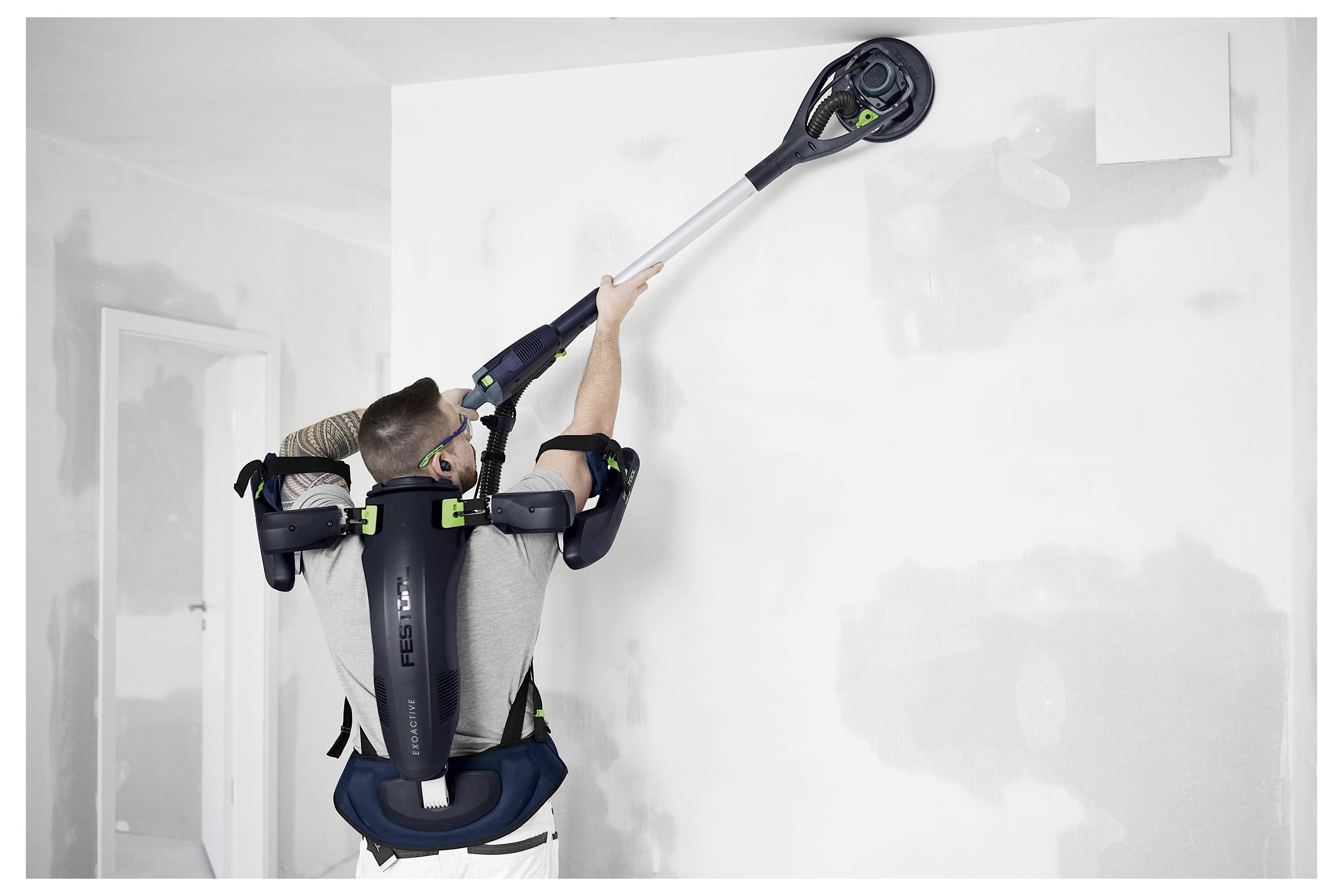 Een persoon die een veiligheidsharnas draagt, gebruikt een elektrisch gereedschap op een muur om oppervlakken in een ruimte onder renovatie glad te maken, wat aangeeft dat het hier om binnenwerk gaat.