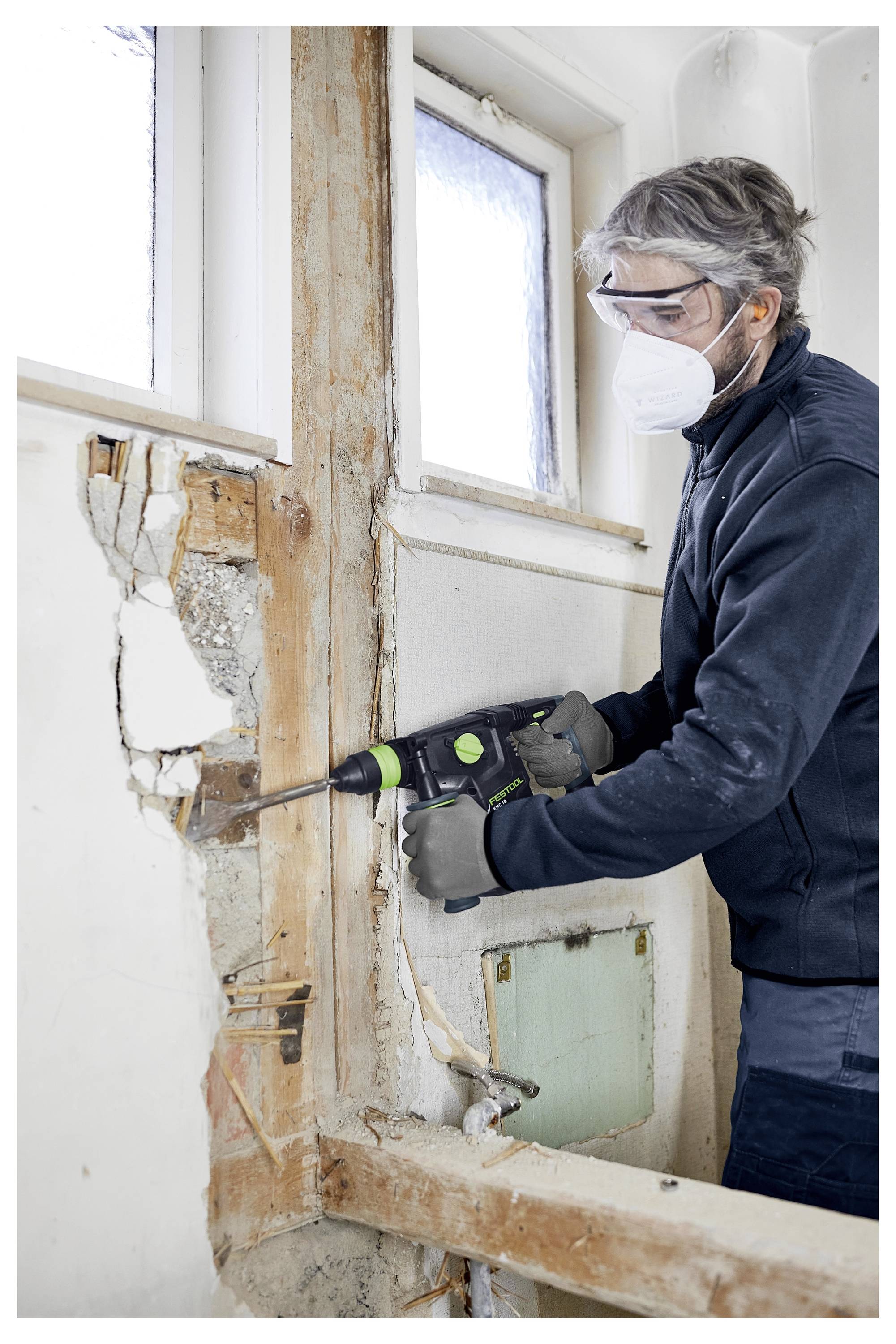 'Persoon met veiligheidsbril en masker gebruikt een elektrisch gereedschap om pleisterwerk van een muur te verwijderen, waardoor een houten en betonnen constructie zichtbaar wordt.'