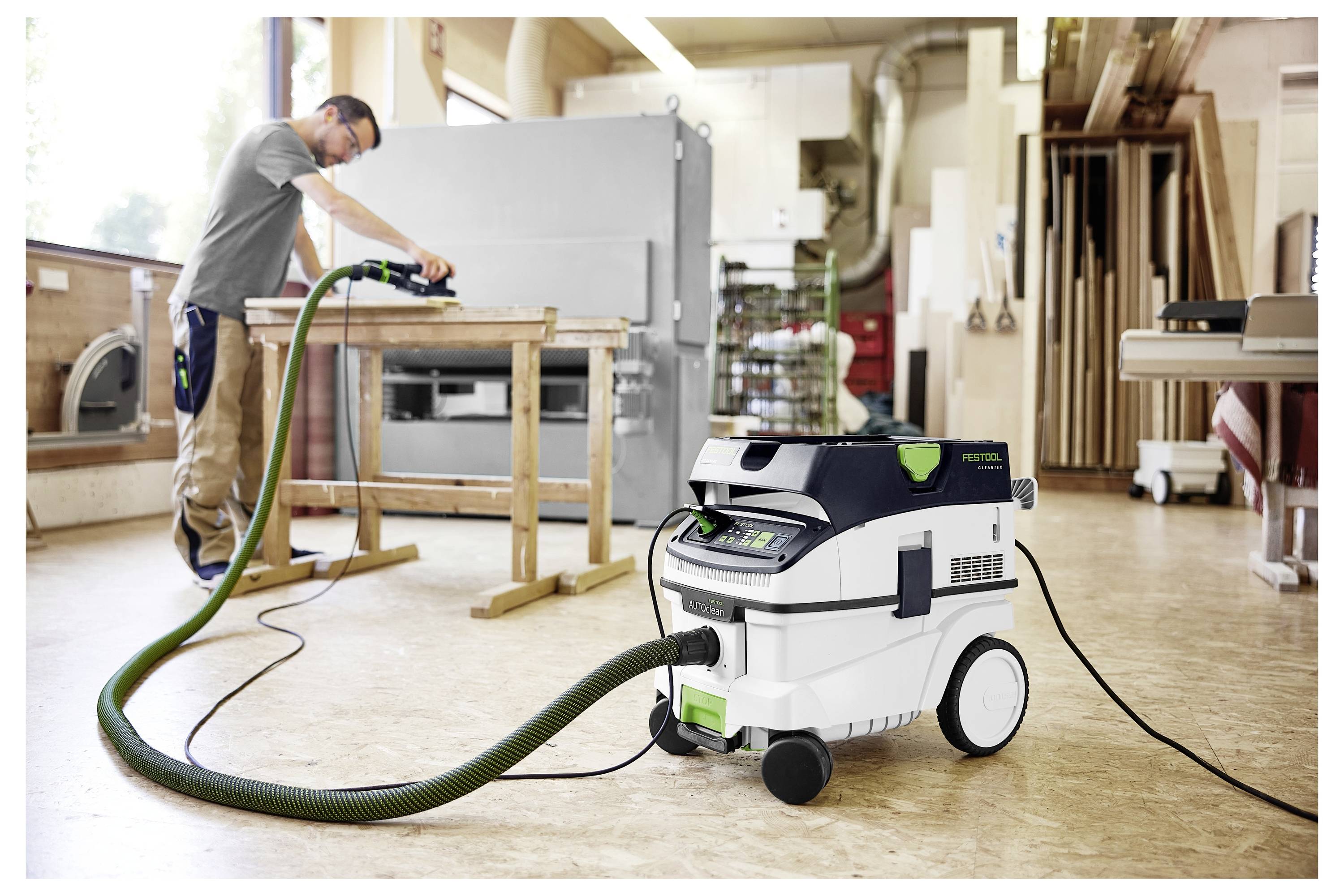 Een persoon gebruikt een elektrisch gereedschap dat is aangesloten op een Festool stofafzuiger in een werkplaats, omringd door houtbewerkingsapparatuur en materialen.