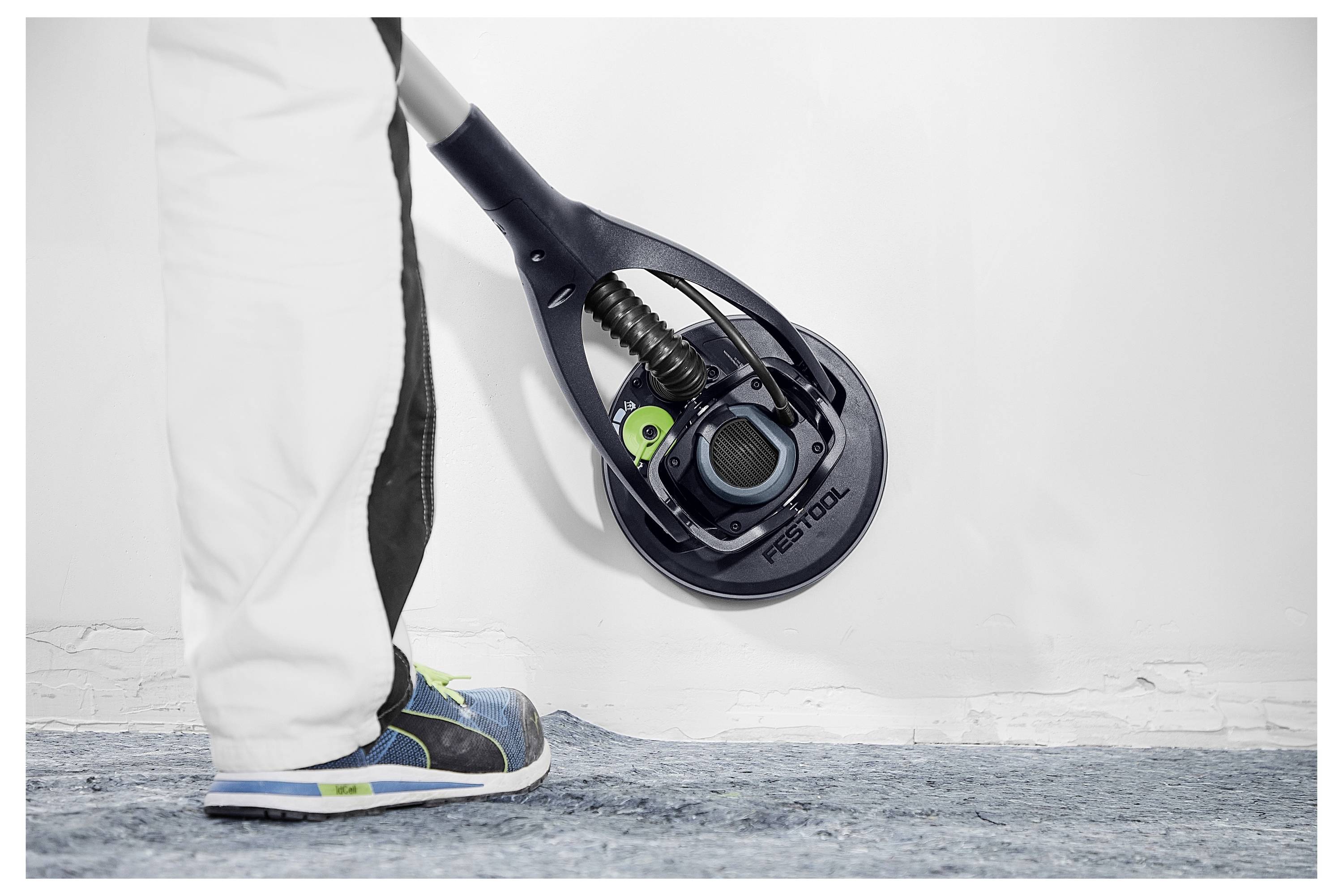 'Persoon die een Festool wandschuurmachine op een wit oppervlak gebruikt, gekleed in witte broek en blauwe sneakers. Close-up van het schuurproces.'