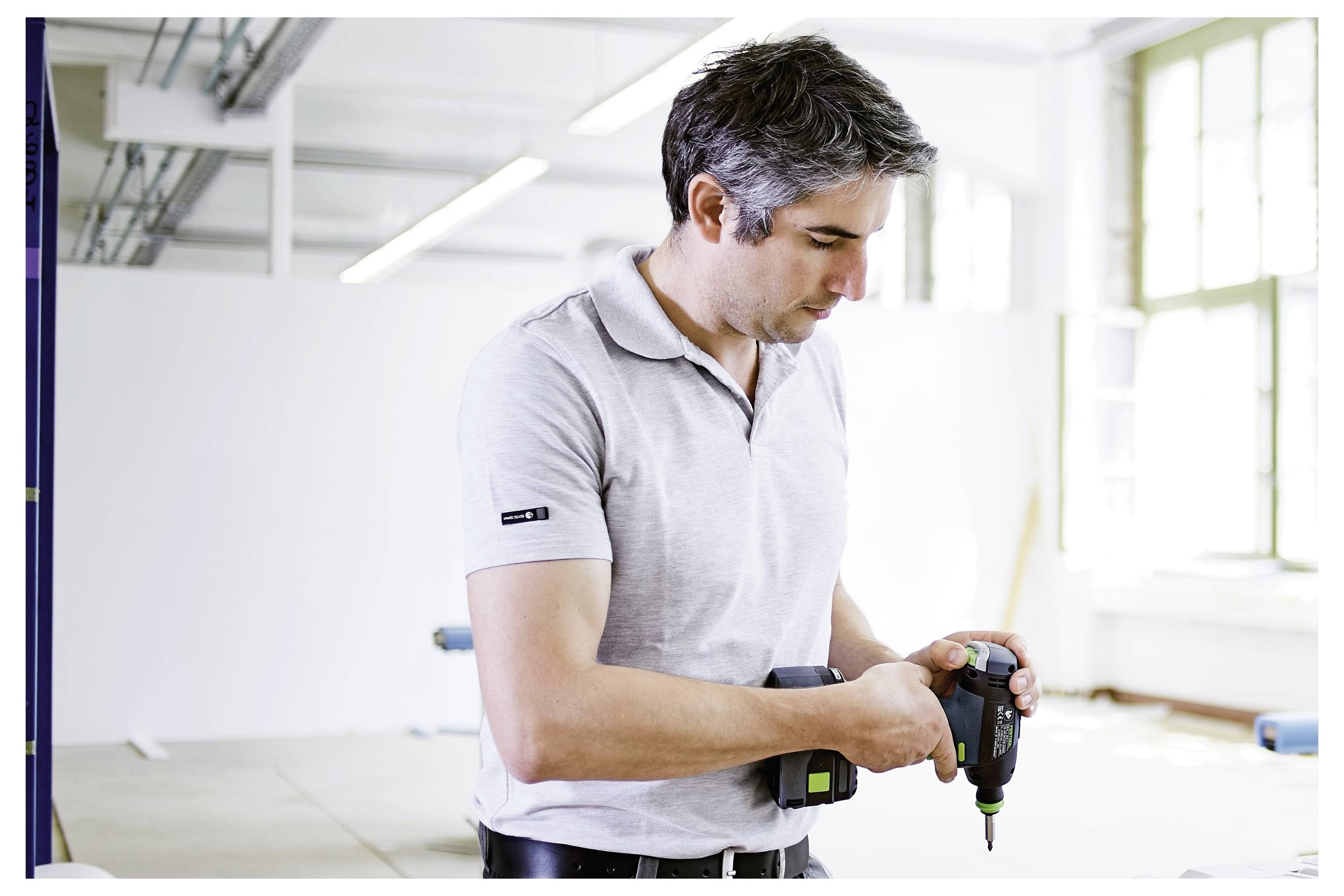 Een man in een lichtgrijs poloshirt gebruikt een elektrische boor in een helderder, ruime werkplaats of studio. De omgeving ziet er industrieel uit met ramen en witte muren.