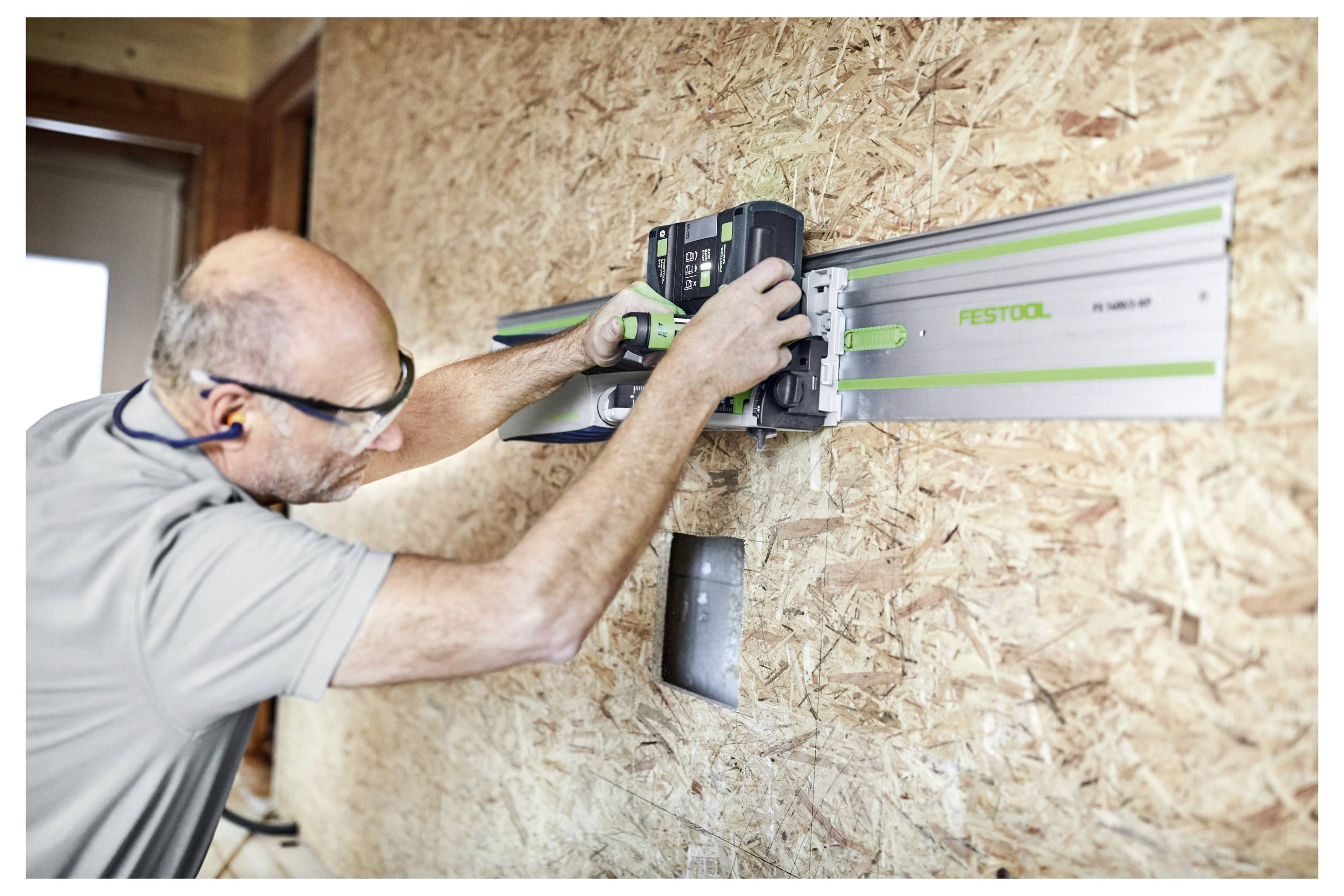 Een persoon die een veiligheidsbril draagt, stelt een gereedschap af op een aan de muur bevestigde geleiderail. De muur is van hout, en een elektrisch stopcontact is zichtbaar eronder.