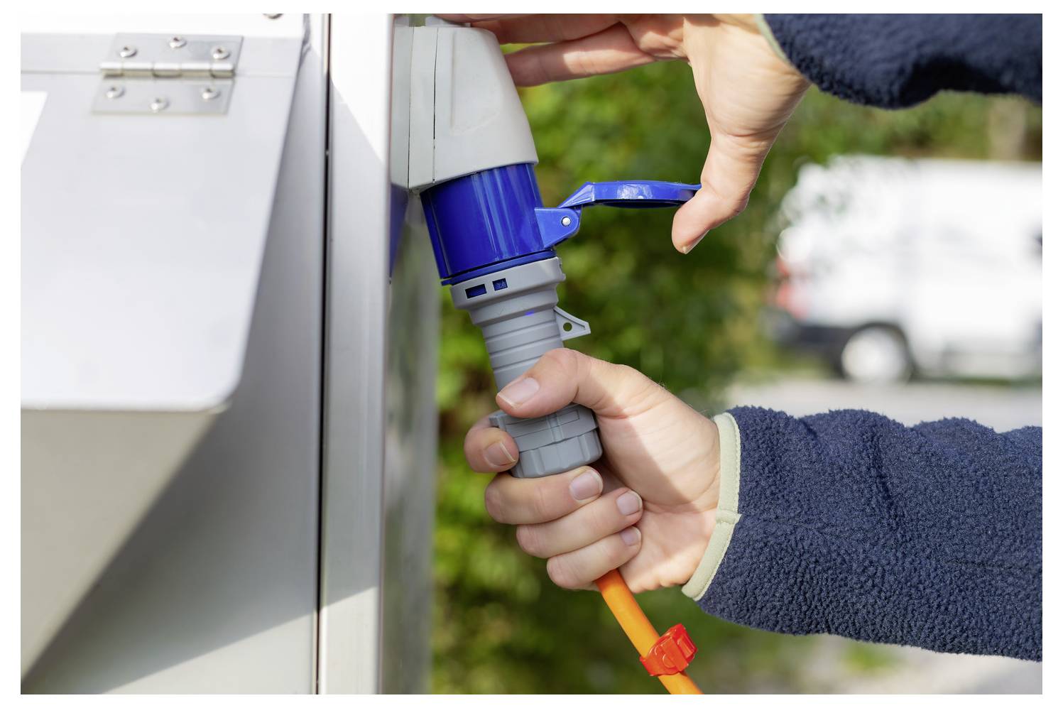 Handen die een oranje stroomkabel in een blauwe buitenelektrische contactdoos op een grijze laadstation steken.
