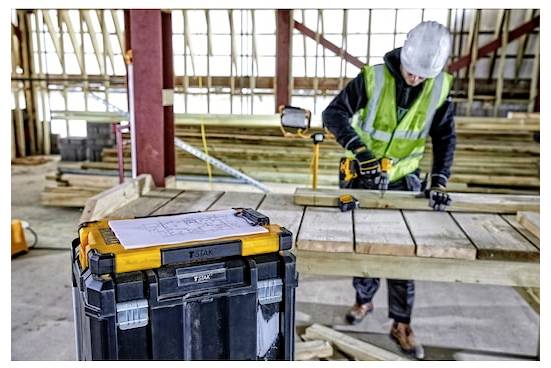 Een bouwvakker in veiligheidsuitrusting gebruikt een boor op houten planken op een bouwplaats. Een gereedschapskist staat op de voorgrond met tekeningen erop.