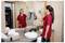 Een vrouw in een rood uniform schoonmaakt een badkamerspiegel met een stoomreiniger. De omgeving is een moderne, nette badkamer met handdoeken die netjes op het aanrecht zijn georganiseerd.