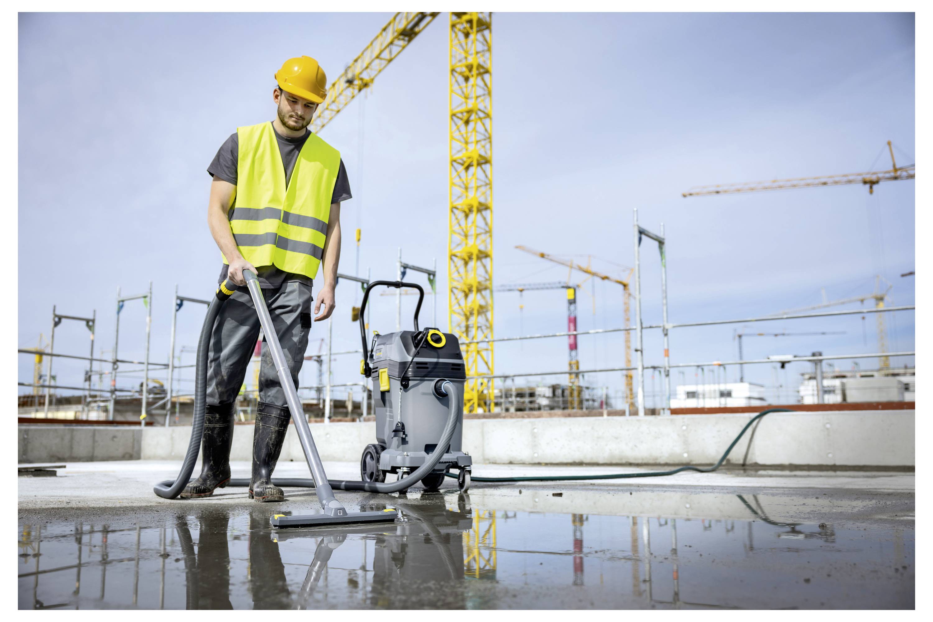 Een bouwvakker in een helm en veiligheidsvest gebruikt een industriële stofzuiger om water op een bouwplaats schoon te maken, met kranen op de achtergrond.