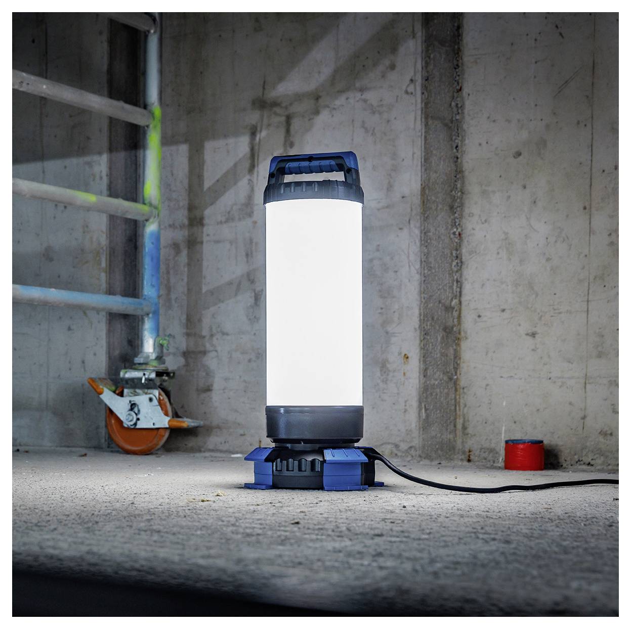 Een helder werklicht op een grijze betonnen vloer naast een ladder en rood object, verlichtend een zwak verlichte industriële ruimte.