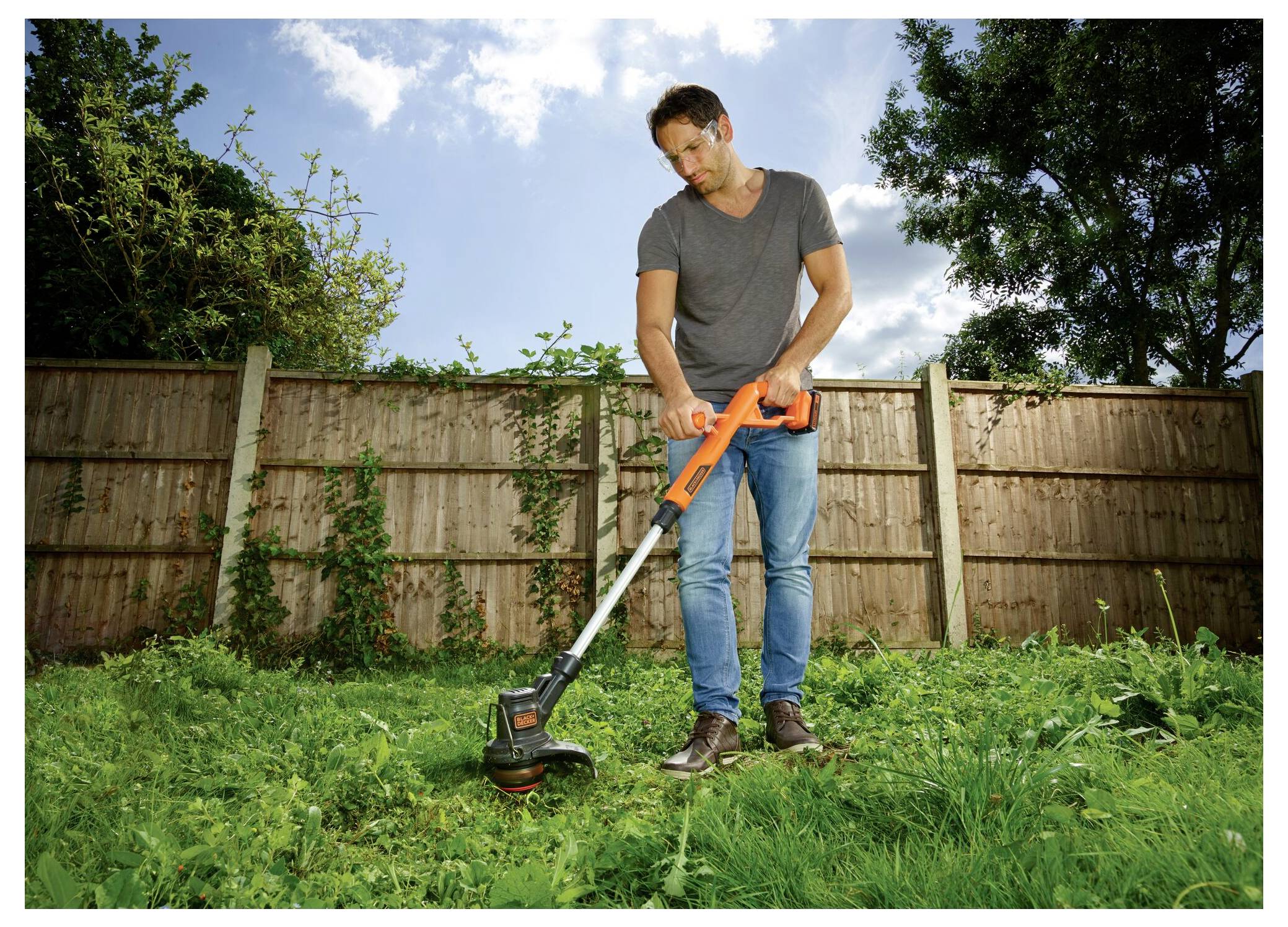 Een persoon gebruikt een oranje grastrimmer om overgroeid gras in een achtertuin te knippen. Een houten hek en bomen zijn zichtbaar op de achtergrond onder een gedeeltelijk bewolkte hemel.