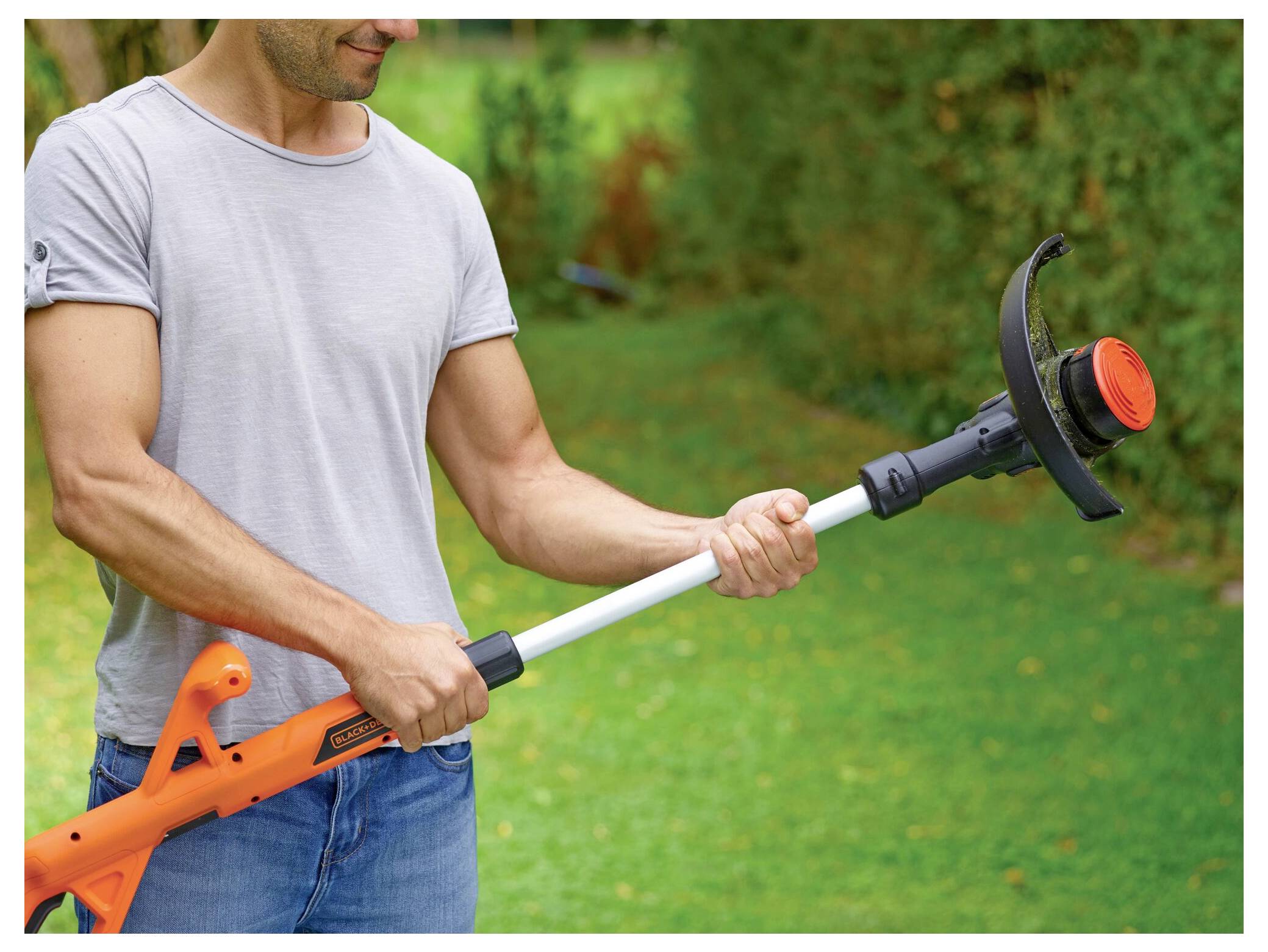 Een persoon in een grijs shirt gebruikt een oranje draadloze grasschaar om gras in een tuin te knippen.