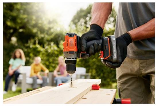 Een persoon met handschoenen gebruikt een elektroboor op een houten plank buiten. Drie personen zitten op een bank in de onscherpe achtergrond.