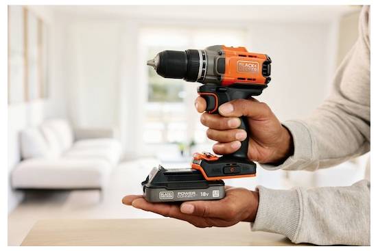 Een persoon houdt een oranje en zwarte draadloze boormachine vast in een helder verlichte ruimte, wat zijn compacte ontwerp en 18V vermogen demonstreert.