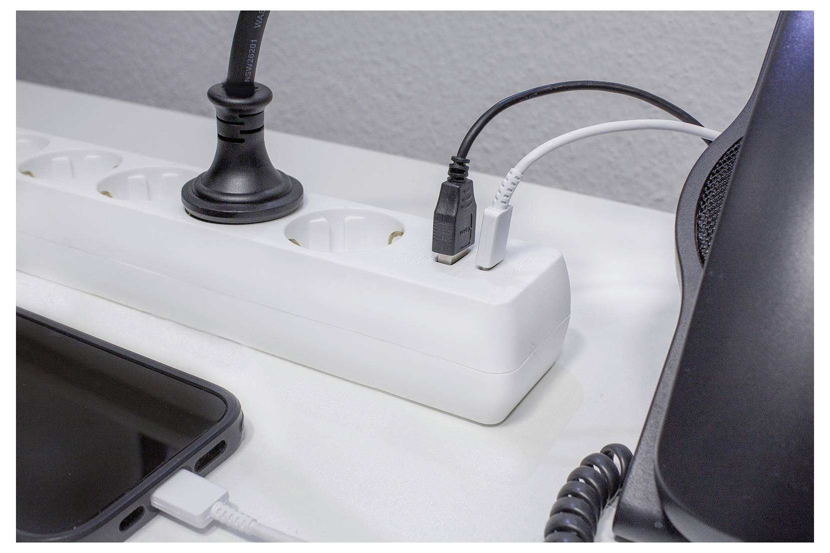 'Een stekkerdoos op een bureau met meerdere stekkers, inclusief een USB-kabel en een telefoonlader. Een smartphone en zwarte luidspreker bevinden zich in de buurt.'