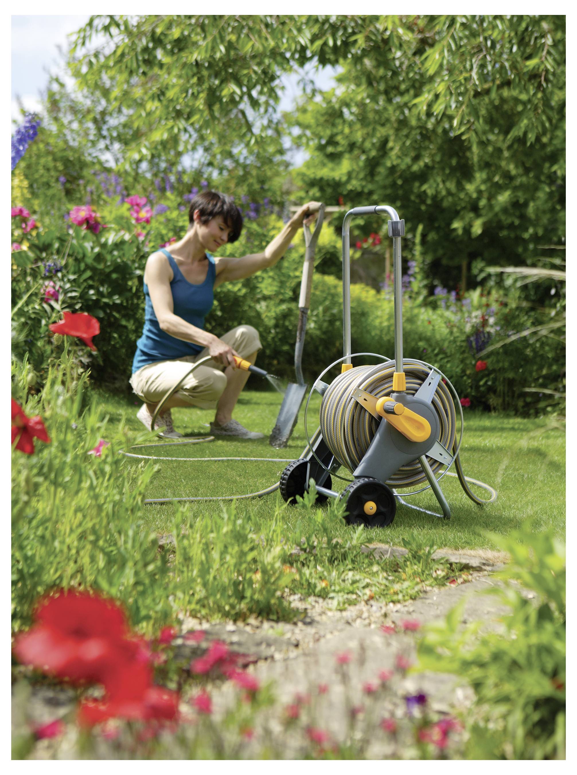 Een persoon die een tuin verzorgt met een slangenhaspel en spade, omgeven door bloeiende bloemen en groen, onder een heldere hemel.