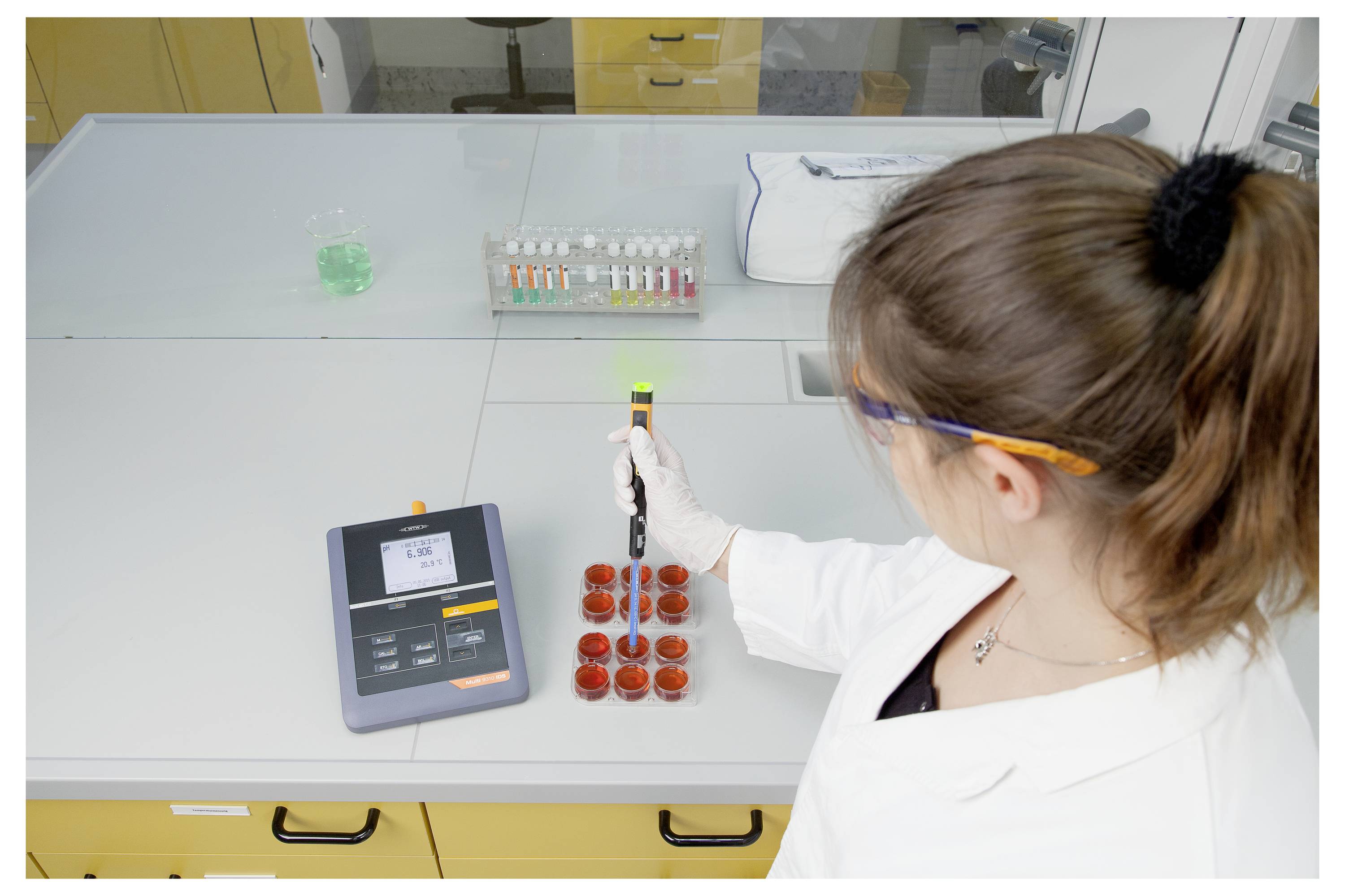Een wetenschapper in een witte jas die een pipet gebruikt op petrischalen die op een laboratoriumtafel staan. Een digitale weegschaal en gekleurde monsters zijn in de buurt zichtbaar.