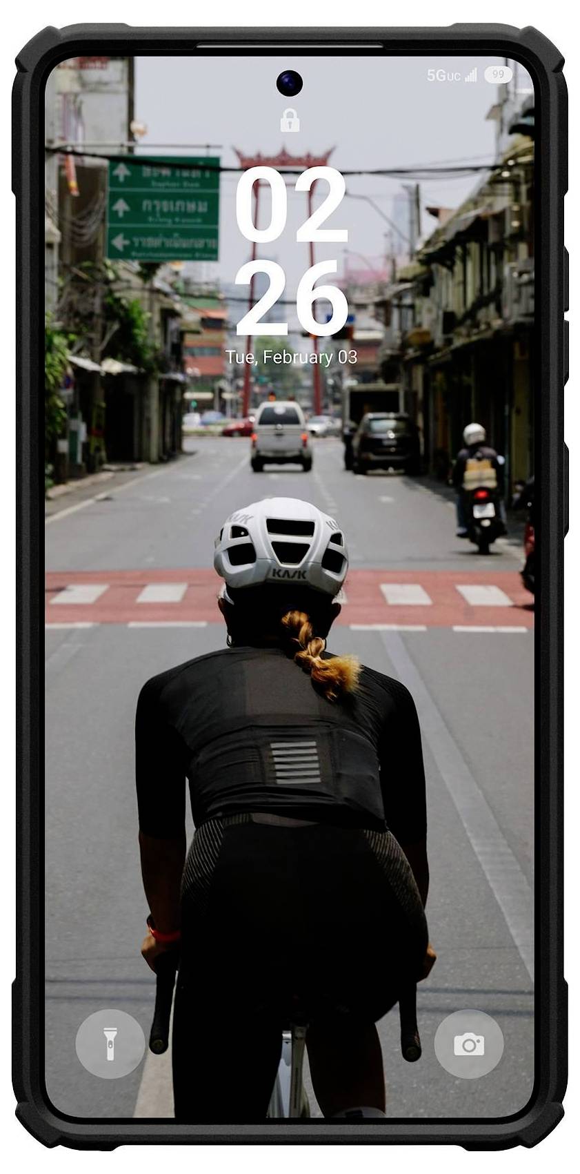 Een persoon op een fiets rijdt een stadsstraat af, gezien van achteren. Het smartphonescherm toont '02:26 PM, din, 3 februari'.