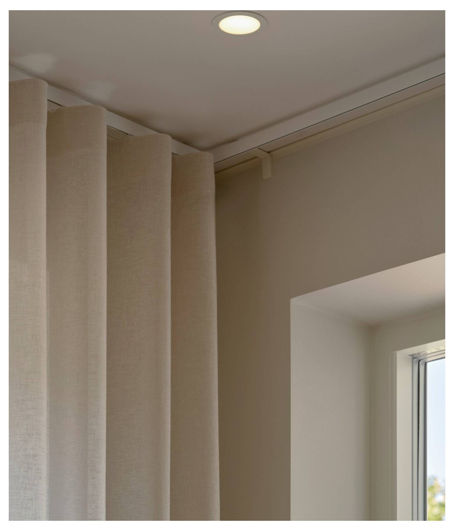 Een beige gordijn dat in de hoek van een kamer naast een raam hangt, met een plafondlamp die aanstaat, wat een gezellige sfeer creëert.