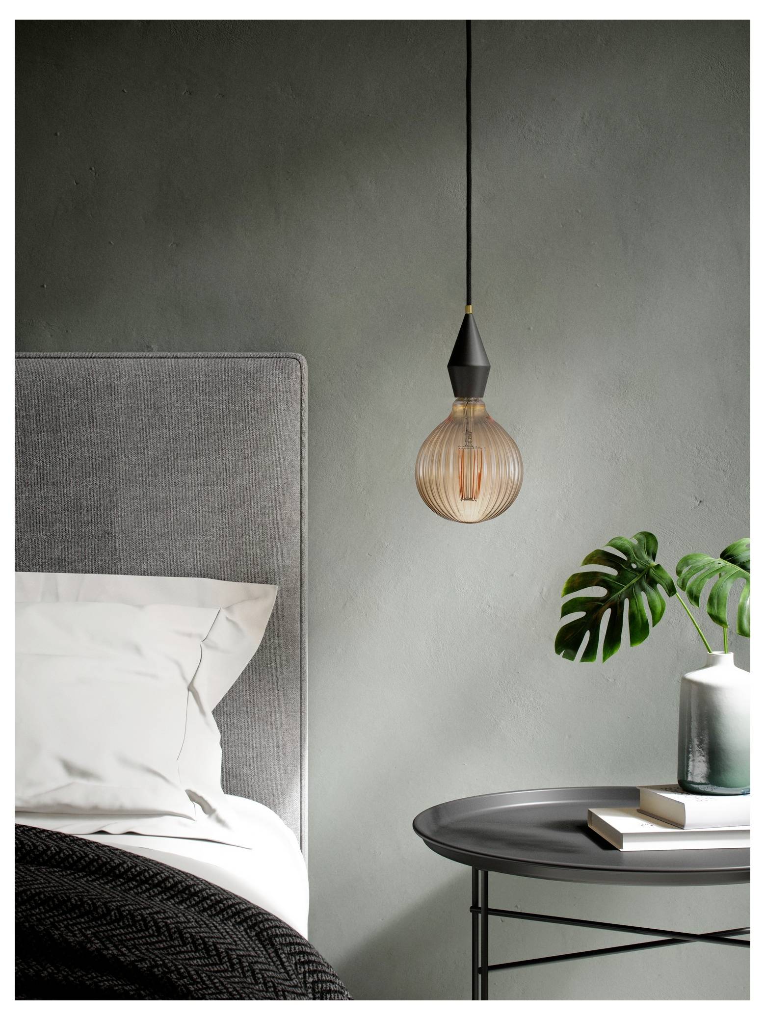 Een modern slaapkamerscène met een hangende hanglamp, een grijs gestoffeerd bed met wit beddengoed, en een klein tafeltje met een plant en boeken.