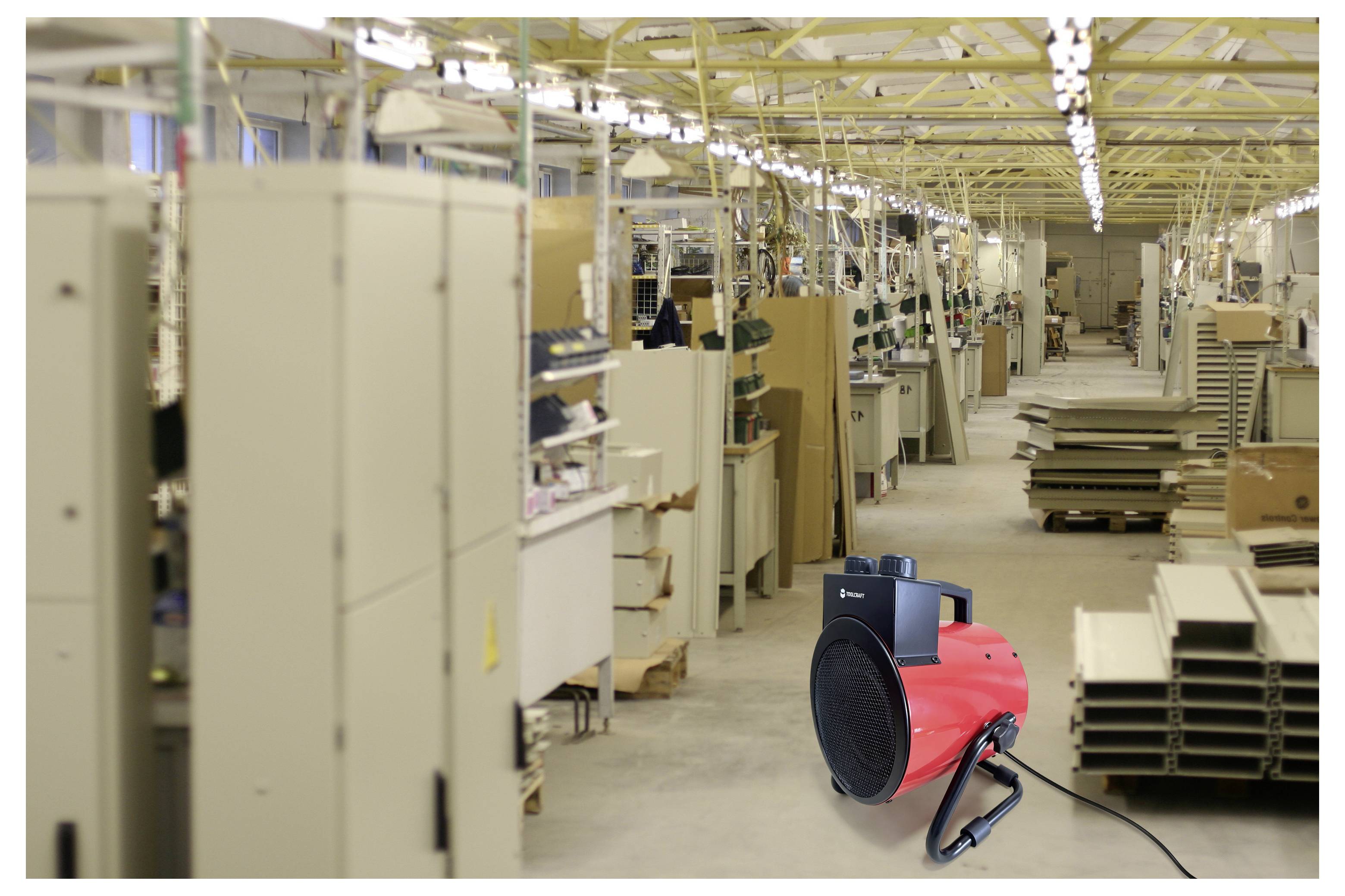 Industriële fabrieksingang met een rode draagbare verwarmer/ventilator op de voorgrond, apparatuur en gestapelde materialen langs de werkplaats.