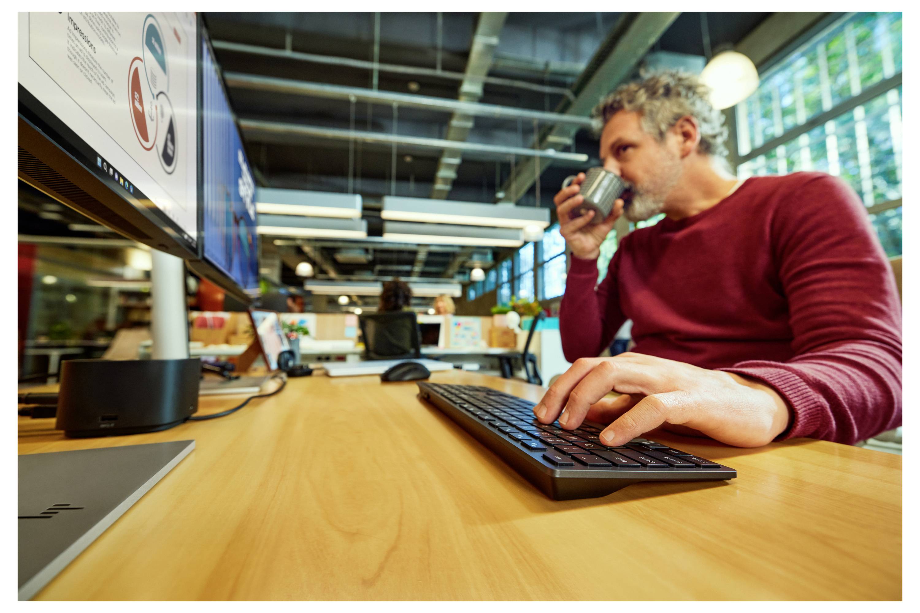 Een man in een rode trui werkt aan een kantoorafdeling, drinkend uit een mok terwijl hij op een toetsenbord tikt, met een computermonitor die grafieken weergeeft.