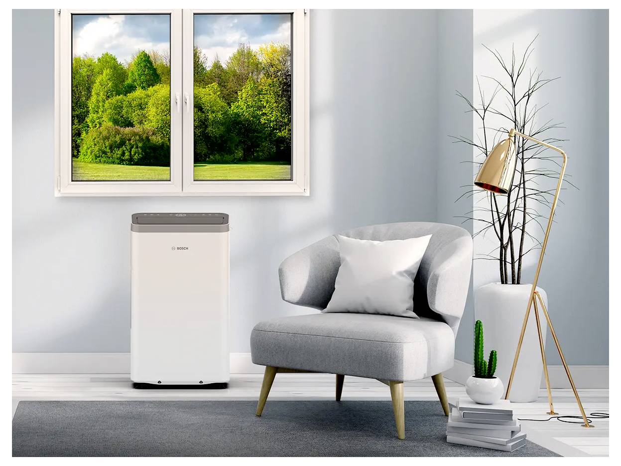 Een woonkamer met een groot raam dat uitzicht biedt op groene bomen, een grijze stoel met een kussen, een draagbare airconditioner en een stijlvolle lamp.