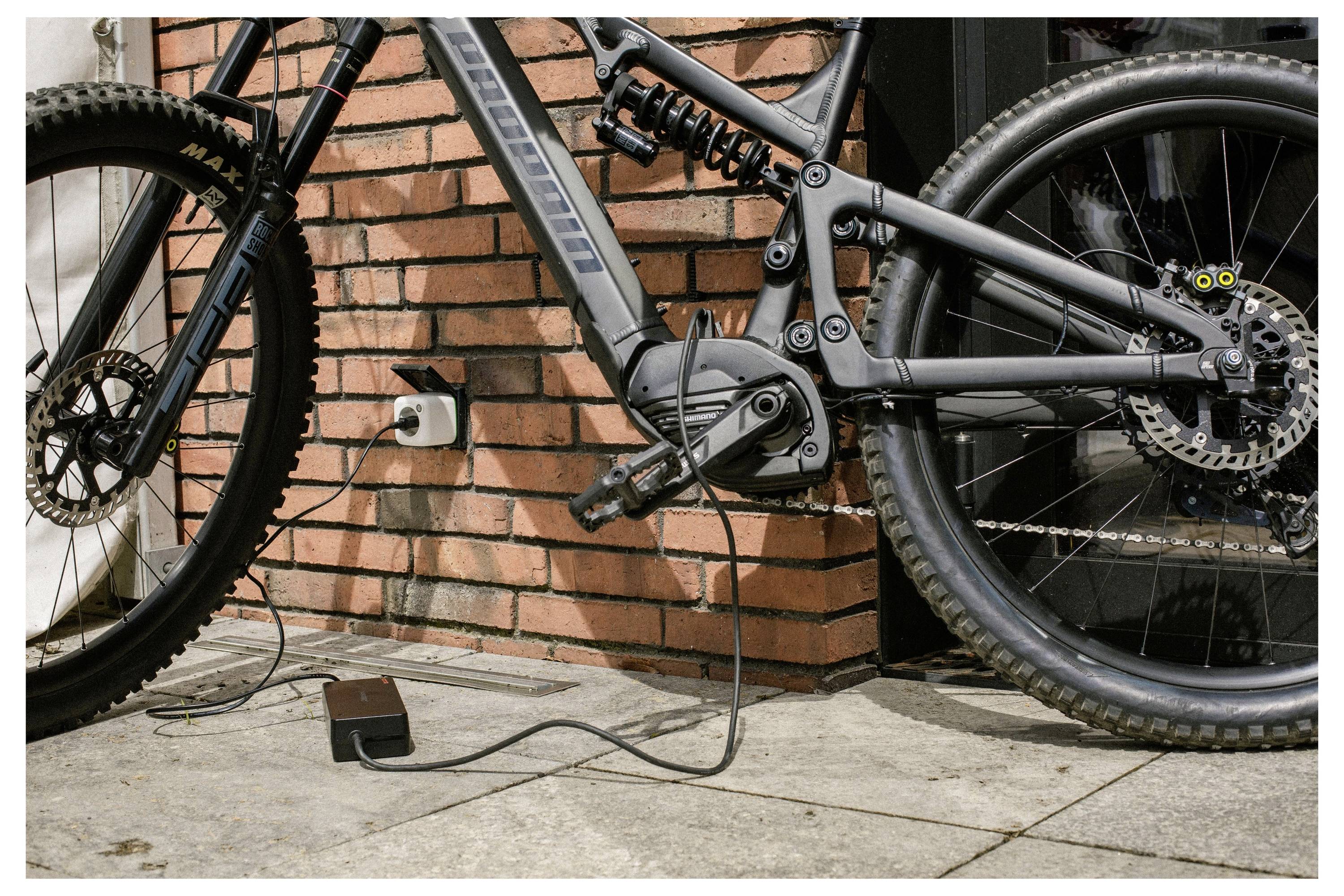 Elektrische fiets aangesloten op een wandcontactdoos, opladen op een patio met bakstenen muur op de achtergrond.