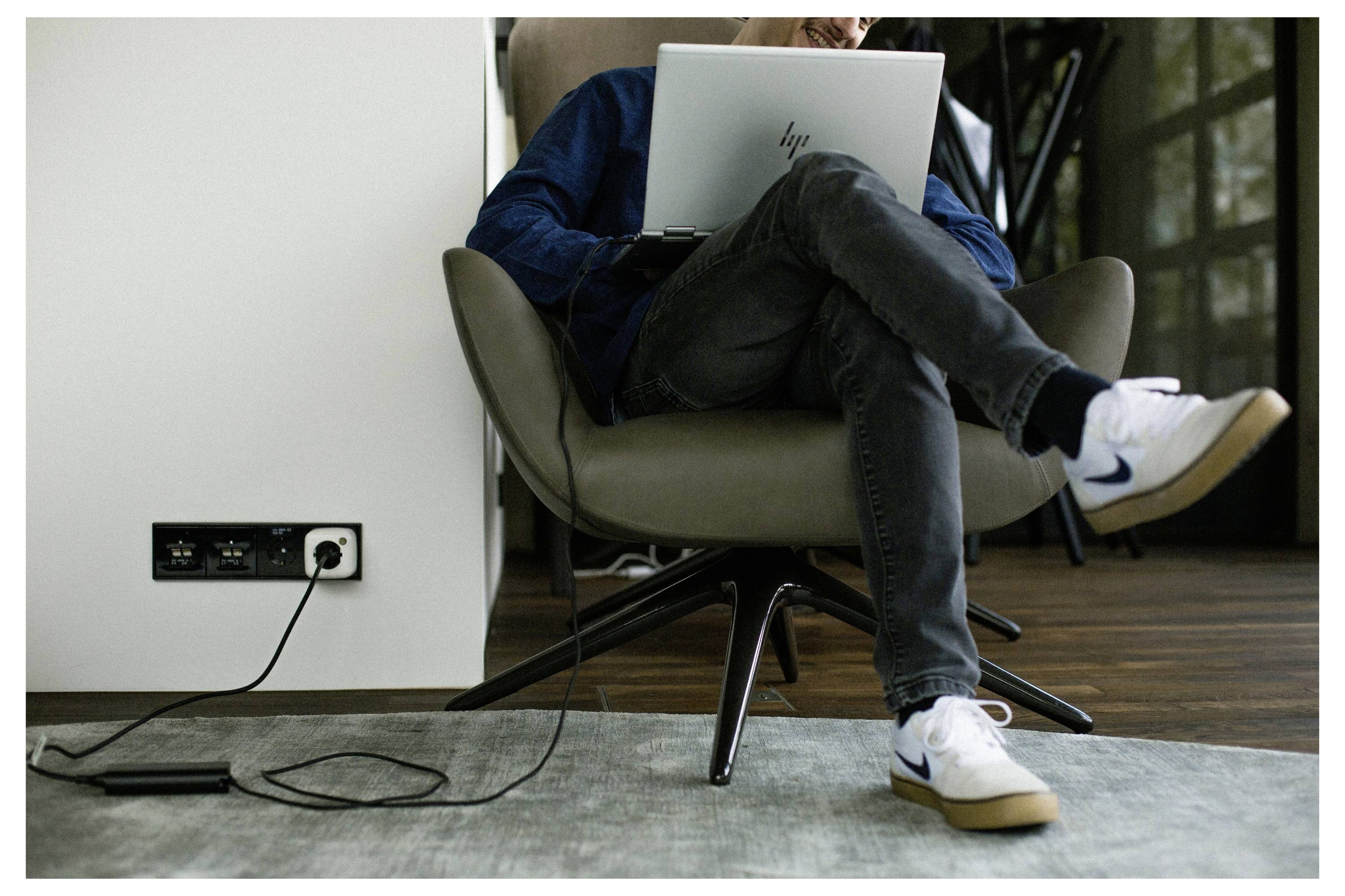 Persoon zittend op een stoel die een laptop gebruikt, met gekruiste benen. Een stroomverdeler ligt op de vloer, aangesloten op een wandcontactdoos. Informele binnenshuis omgeving.