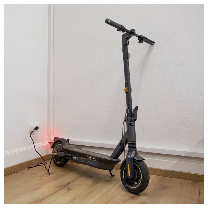 Een elektrische scooter staat binnenshuis opgeladen met een zichtbare rode lampindicator. Het staat tegen een witte muur op een houten vloer.