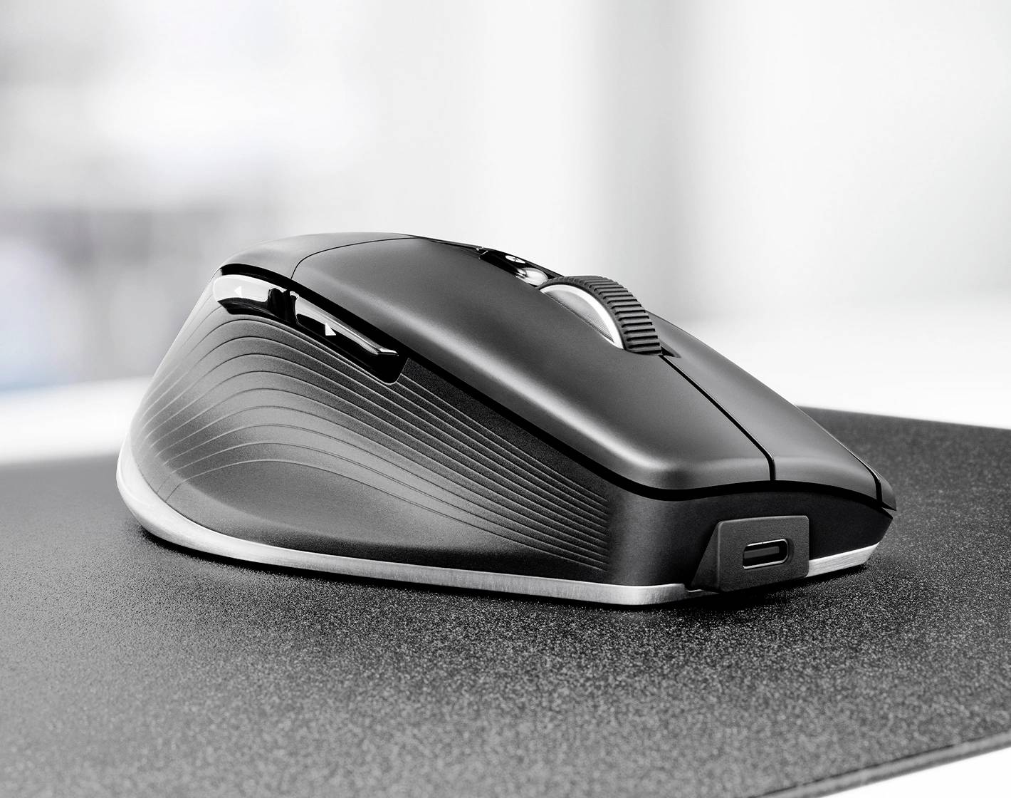 Een slanke, ergonomische zwarte computermuis met een gestructureerde zijgreep rust op een grijze muismat, met een zichtbare USB-oplaadpoort.