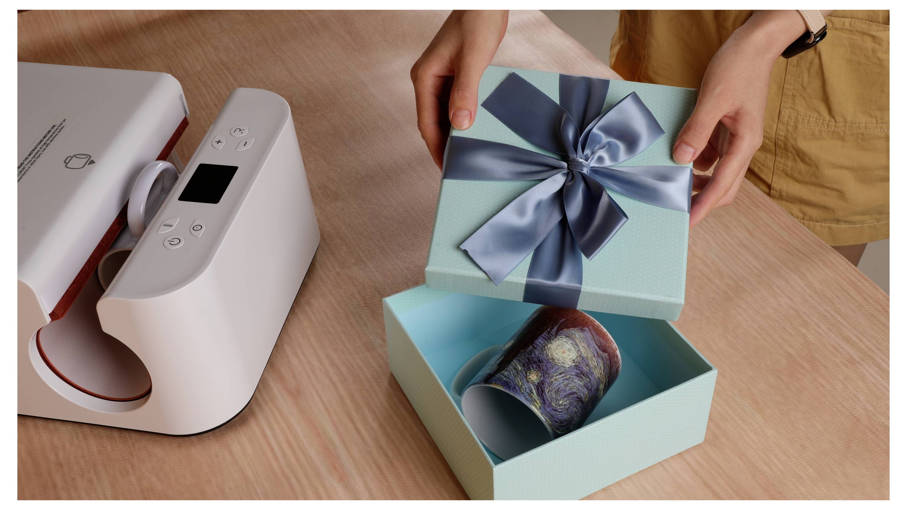 'Een persoon opent een tealkeurige geschenkdoos met een blauw lint, waaruit een mok tevoorschijn komt, geplaatst op een tafel naast een warmtepersmachine.'
