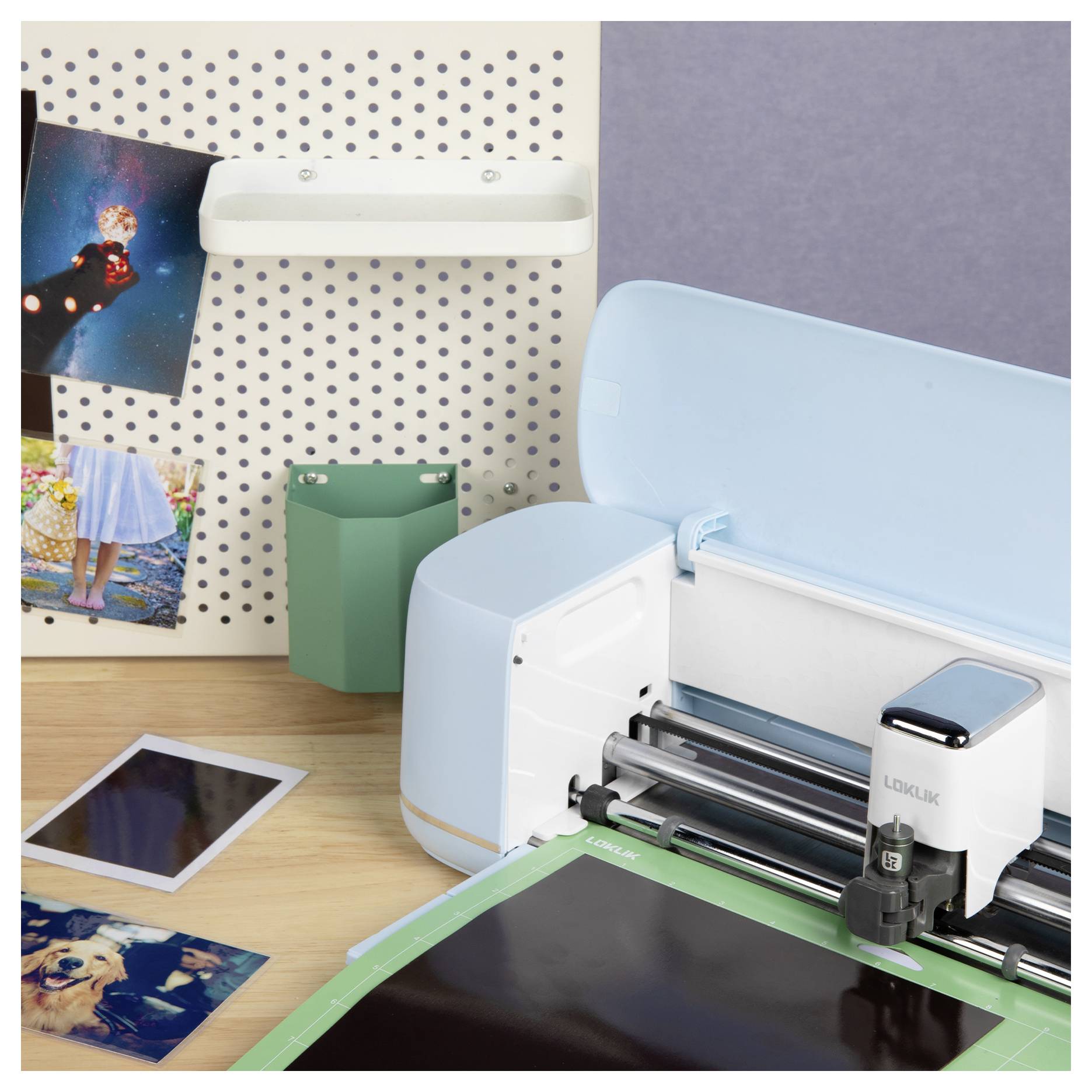 Een snijmachine op een tafel naast foto's en craftmaterialen, met een wandpegboard waarop meer foto's zijn uitgestald en een penhouder.