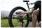 Een persoon op een groene mountainbike controleert de bandenspanning van het achterwiel met een digitale manometer buiten op een grasppad, met heuvels op de achtergrond.