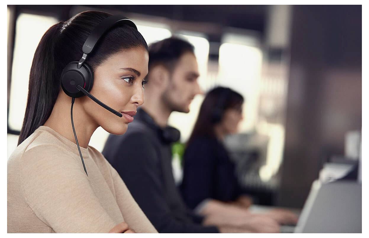 Een vrouw met een headset werkt aan een computer in een callcenter-omgeving. Twee andere personen zijn in de achtergrond wazig.