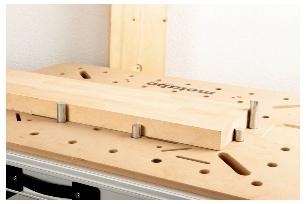 Een houten werkbank met een beige oppervlak bevat een rechthoekige houten plank die op zijn plaats wordt gehouden door cilindrische pennen. Het merk 'Mafell' is zichtbaar.