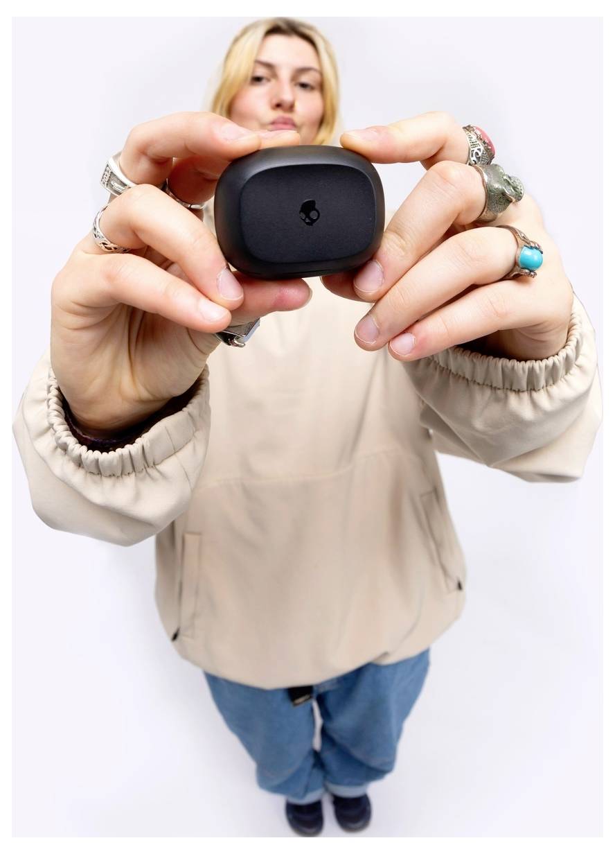 Een persoon in een beige jas en blauwe broek houdt een klein zwart rechthoekig apparaat naar de camera gericht, waardoor de aandacht op het apparaat wordt gevestigd.