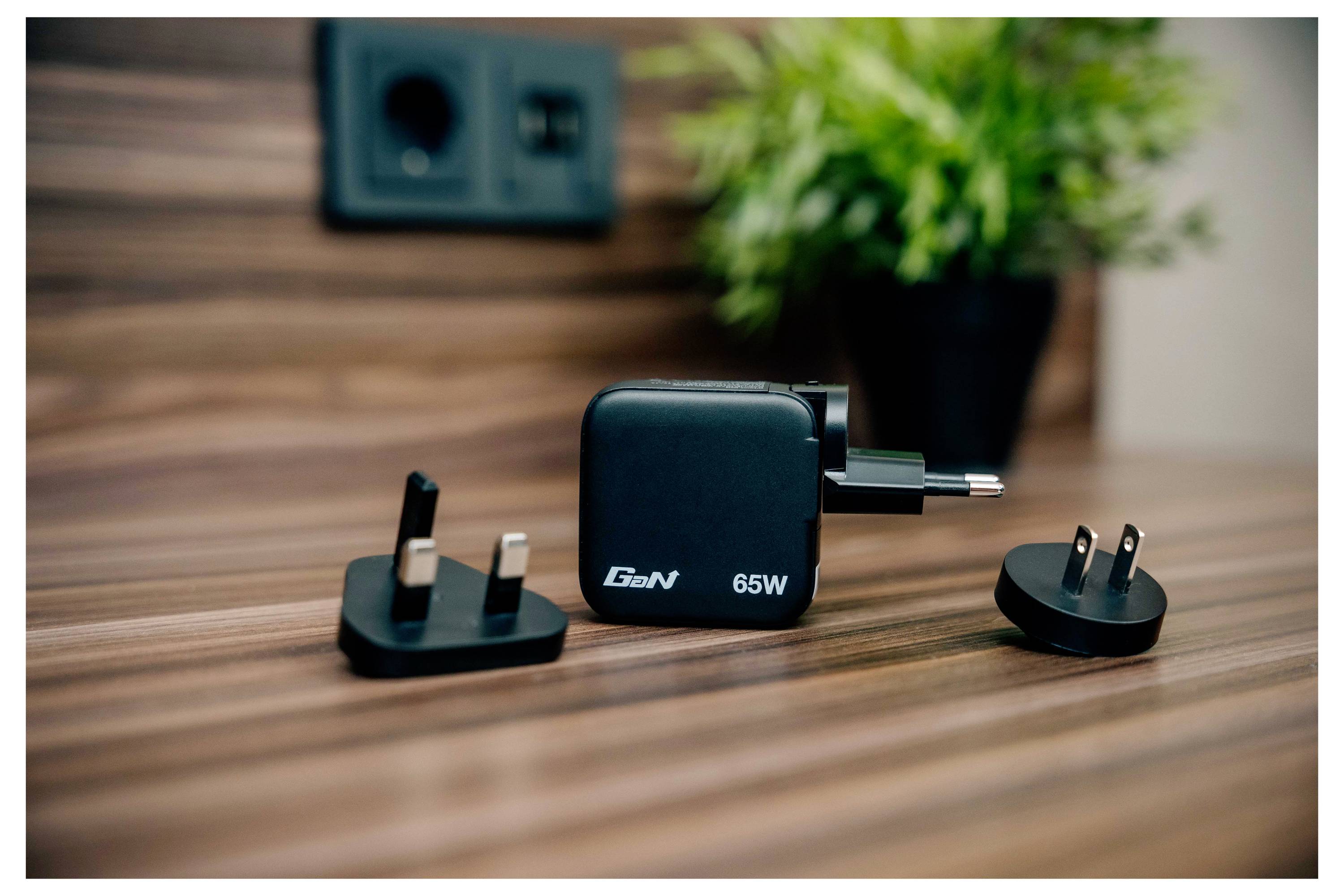 Een zwarte GaN 65W-lader met drie verwisselbare stekkeradapters staat op een houten oppervlak. Een wandcontactdoos en potplant staan op de achtergrond.
